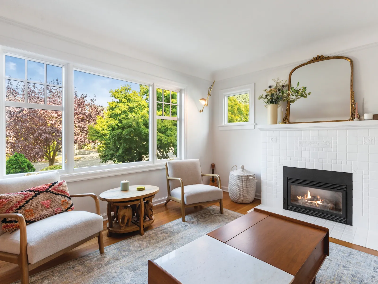Bright living room with large windows, two beige armchairs, wooden coffee table, and a white tiled fireplace.