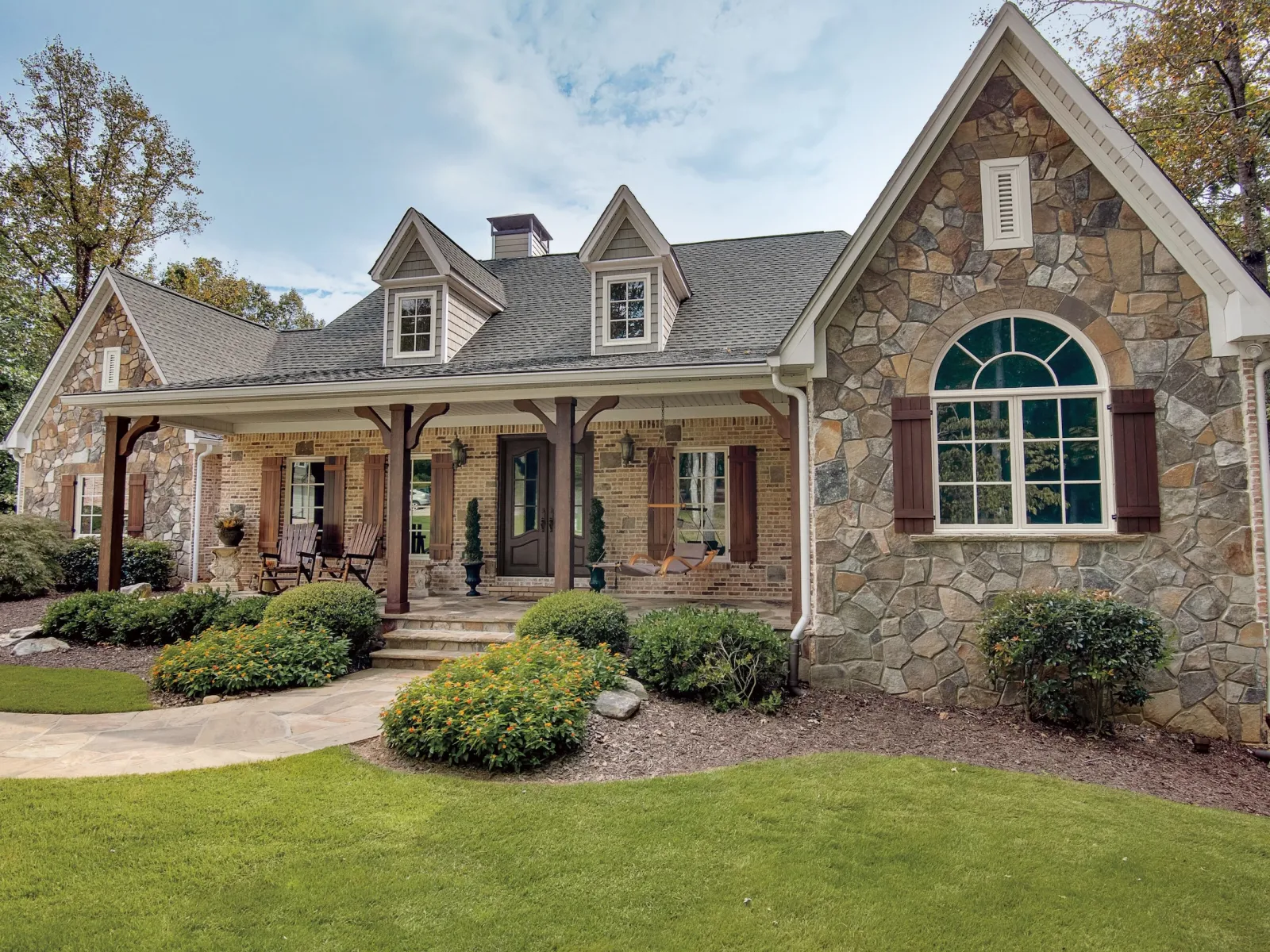 Stone and brick exterior country house with porch, landscaped garden, and lush green lawn under a cloudy sky