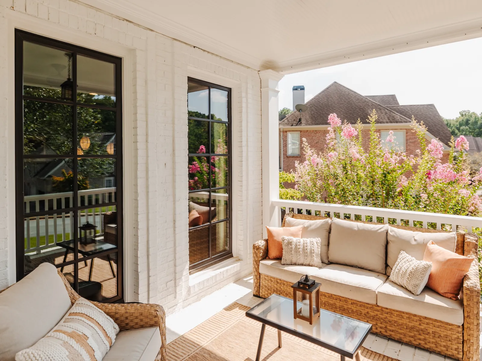 Cozy outdoor patio with wicker sofa, neutral cushions, glass coffee table, and blooming pink flowers in the background.