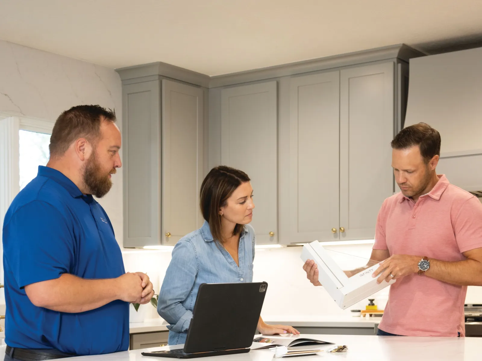 Three adults discuss kitchen renovation plans with samples and laptop in modern kitchen with gray cabinets.