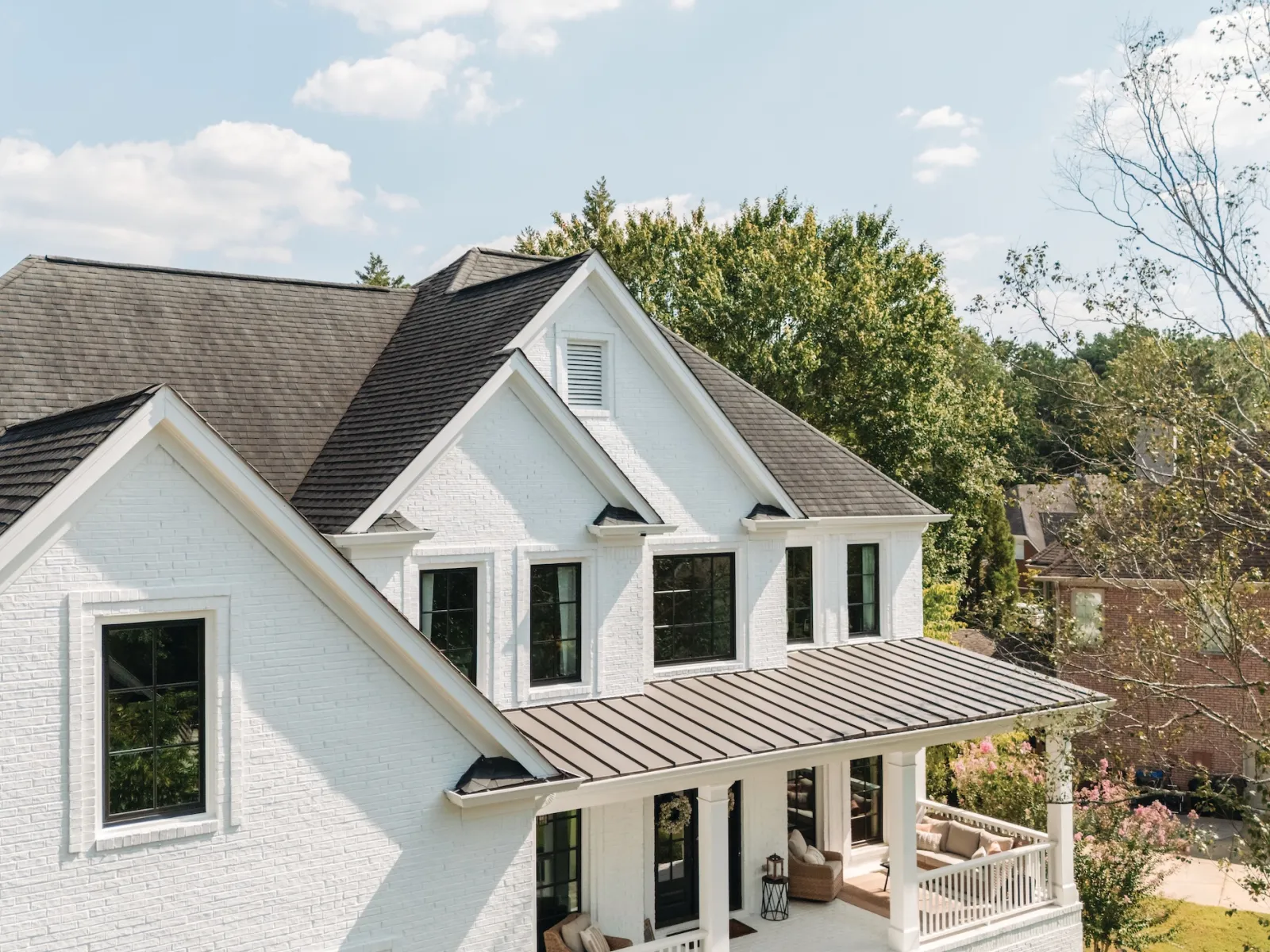 White two-story house with black roof, large windows, and a covered front porch surrounded by trees under a partly cloudy sky