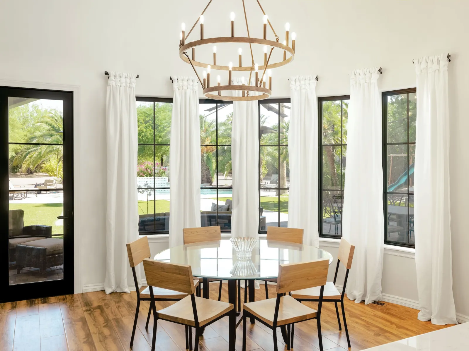 Bright dining area with round glass table, six wooden chairs, chandelier, large windows, and white curtains overlooking a pool.