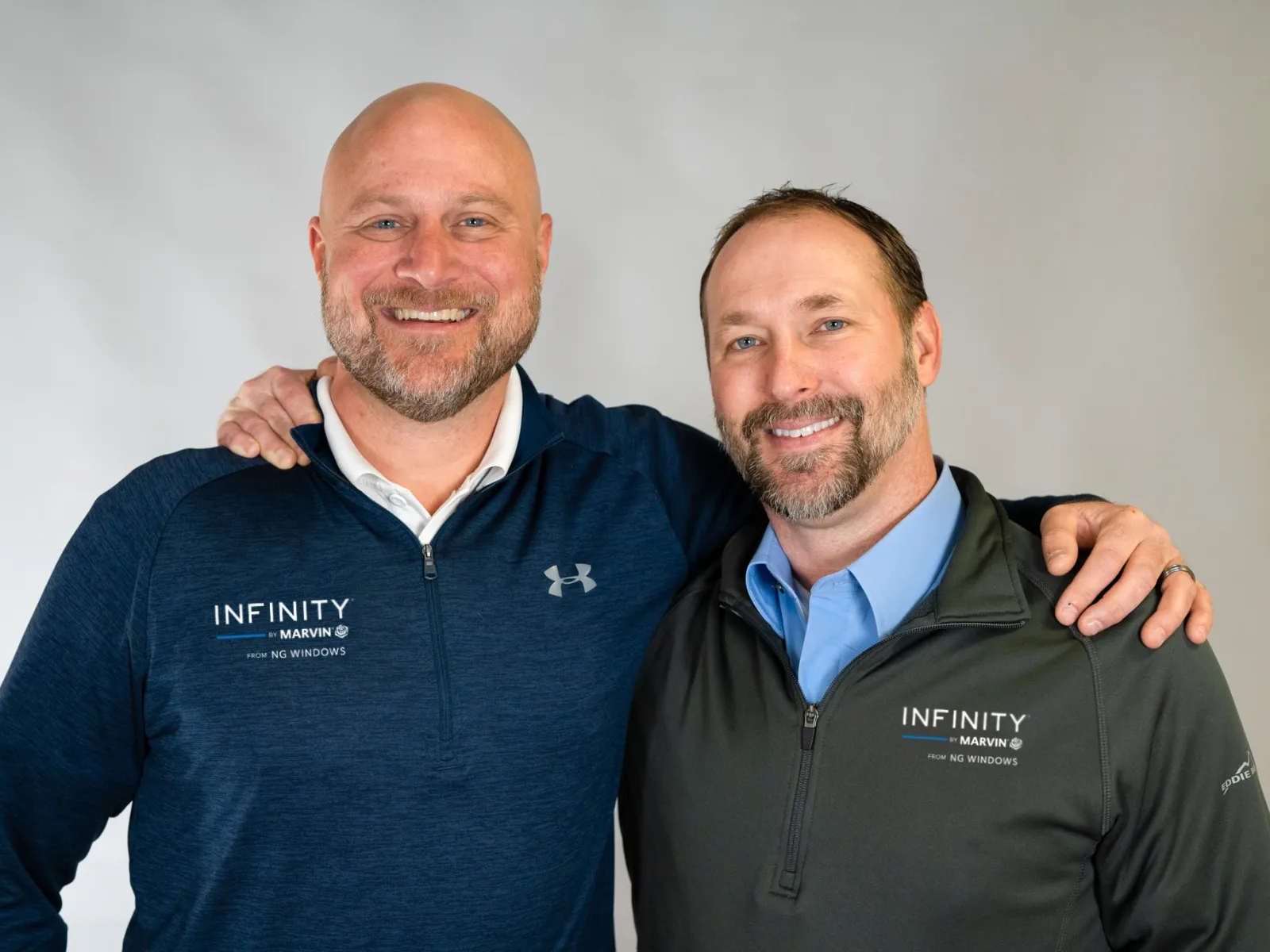 Two men smiling with arms around each other wearing Infinity by Marvin branded jackets against a white background