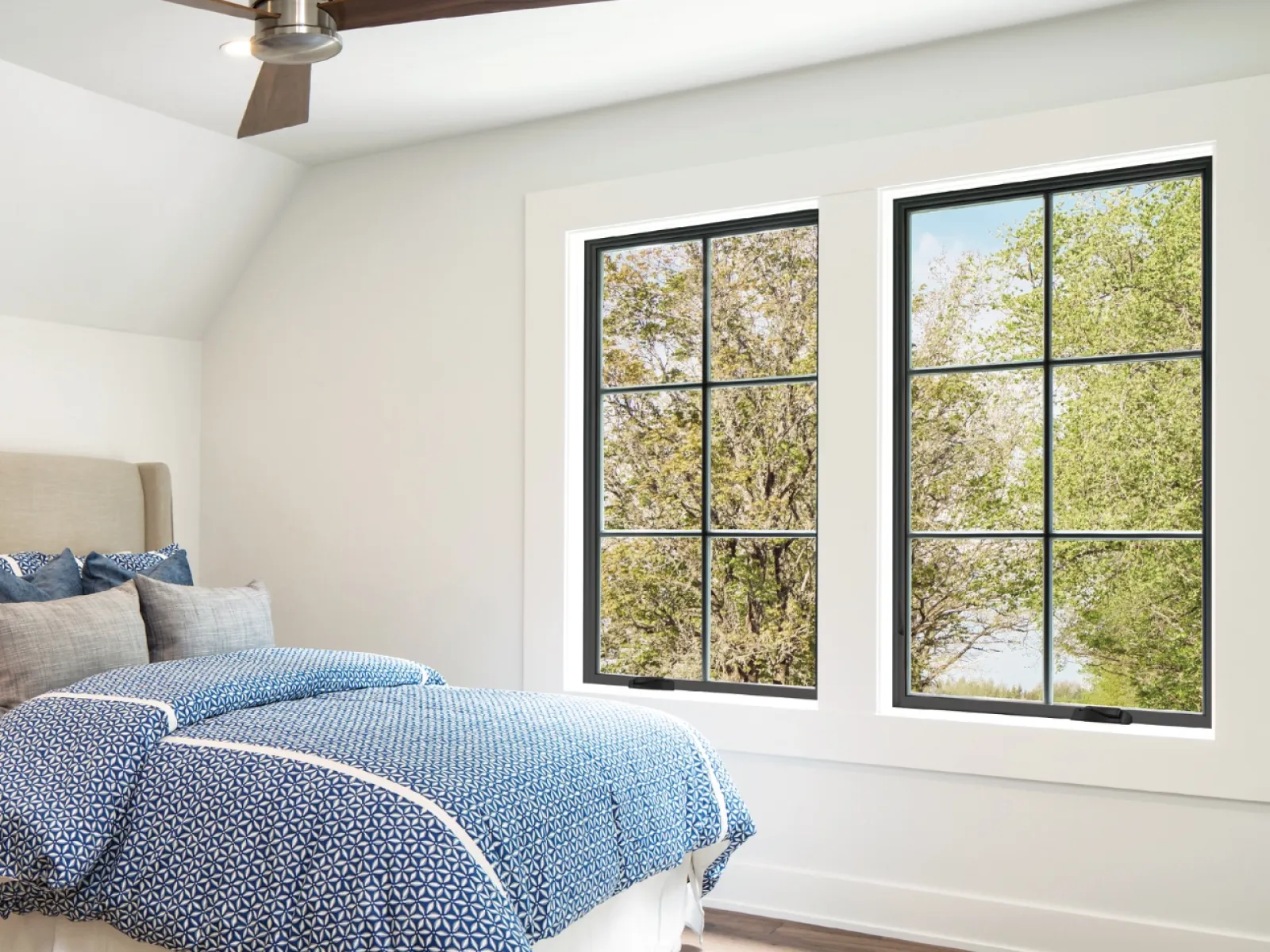A bedroom with ceiling fan, bed with blue comforter, and 2 black casement windows with simulated divided lites overlooking the trees outside