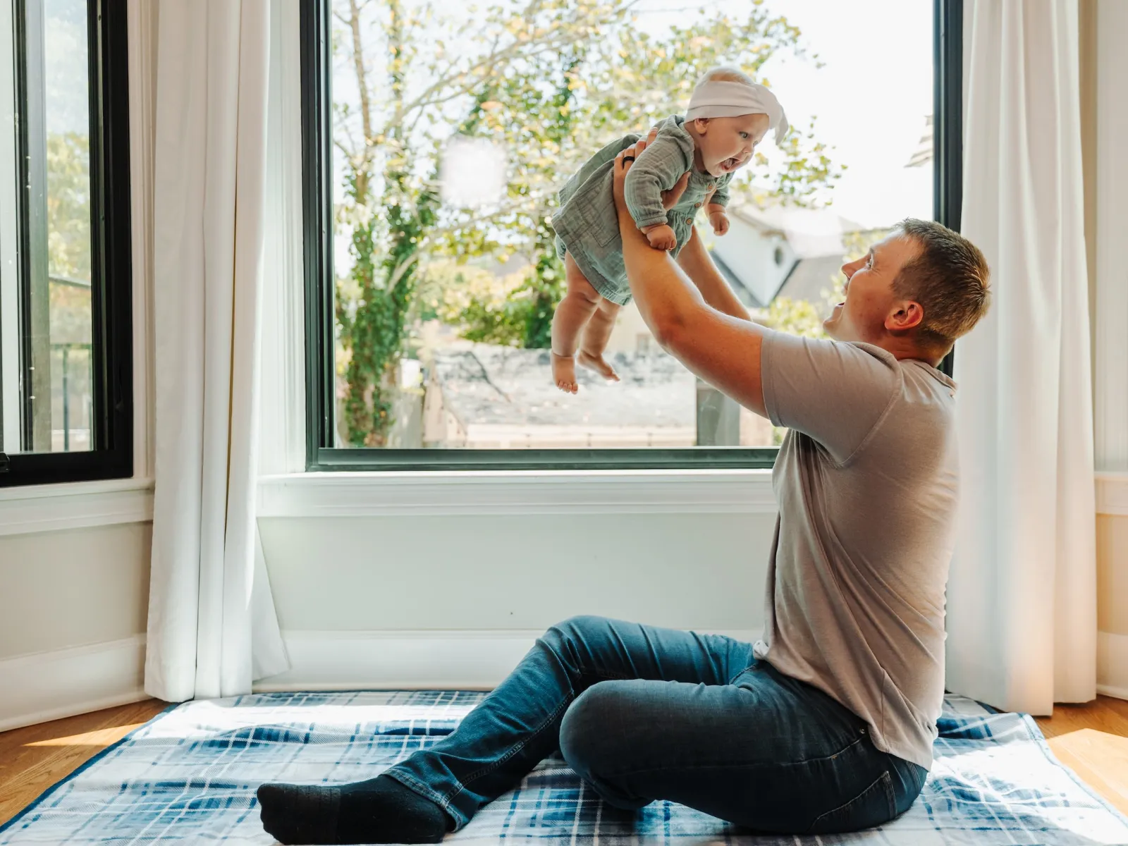 Father joyfully lifts baby indoors by large windows with green trees outside on a sunny day.