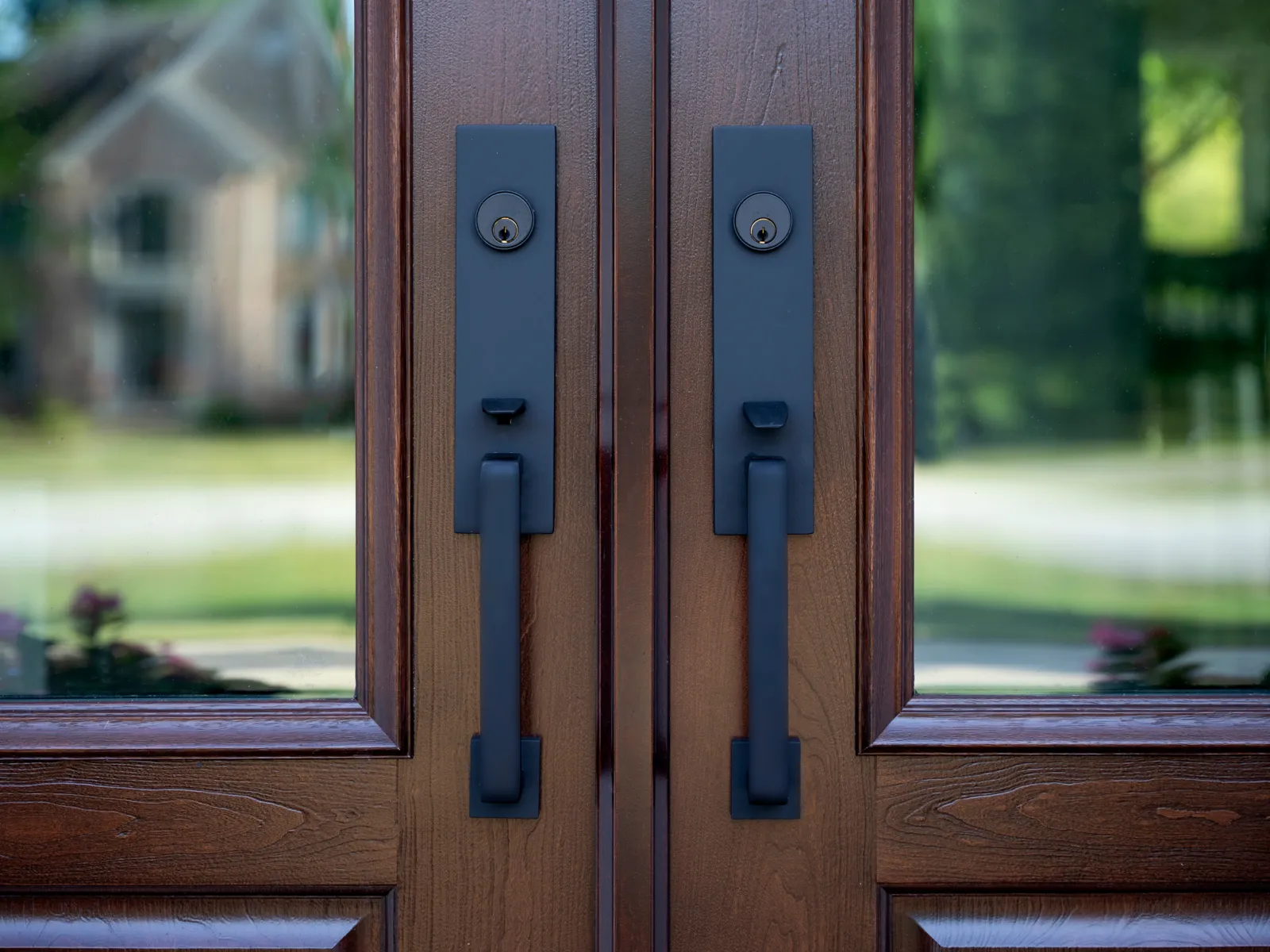 Close-up of wooden double doors with black metal handles and locks reflecting a suburban house and greenery.