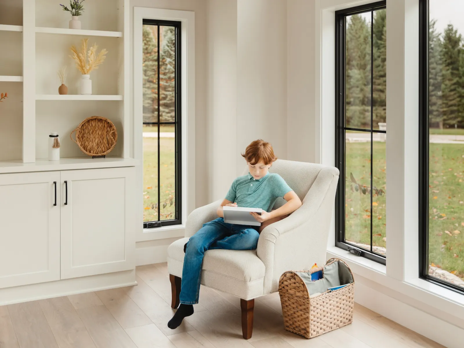 Young boy sitting in a beige armchair reading a book in front of large black casement fiberglass windows.