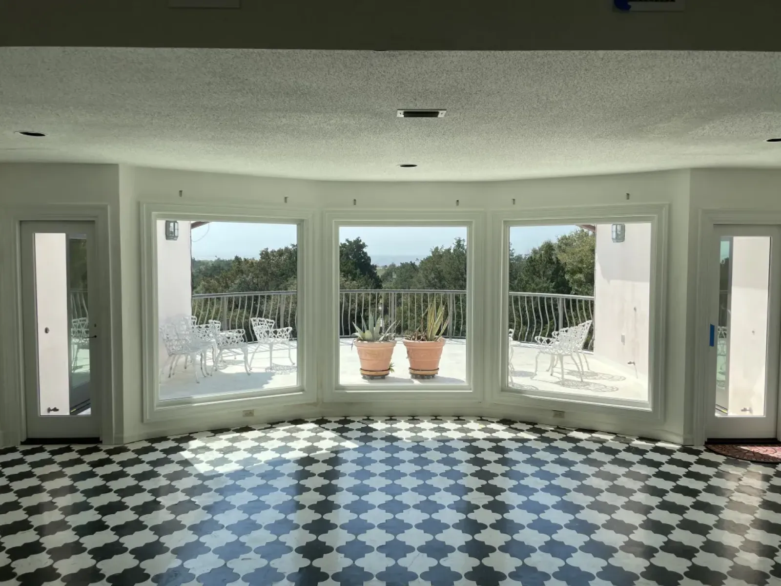 Bright room with black and white tiled floor, three large windows, and outdoor patio with white chairs and potted plants.