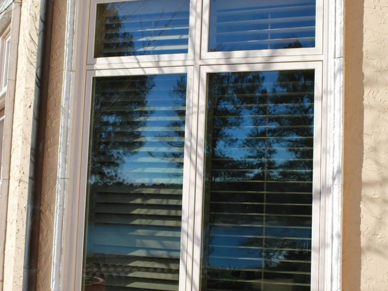 Large window with white frame and shutters reflecting trees and sky on a beige stucco wall with stone base.
