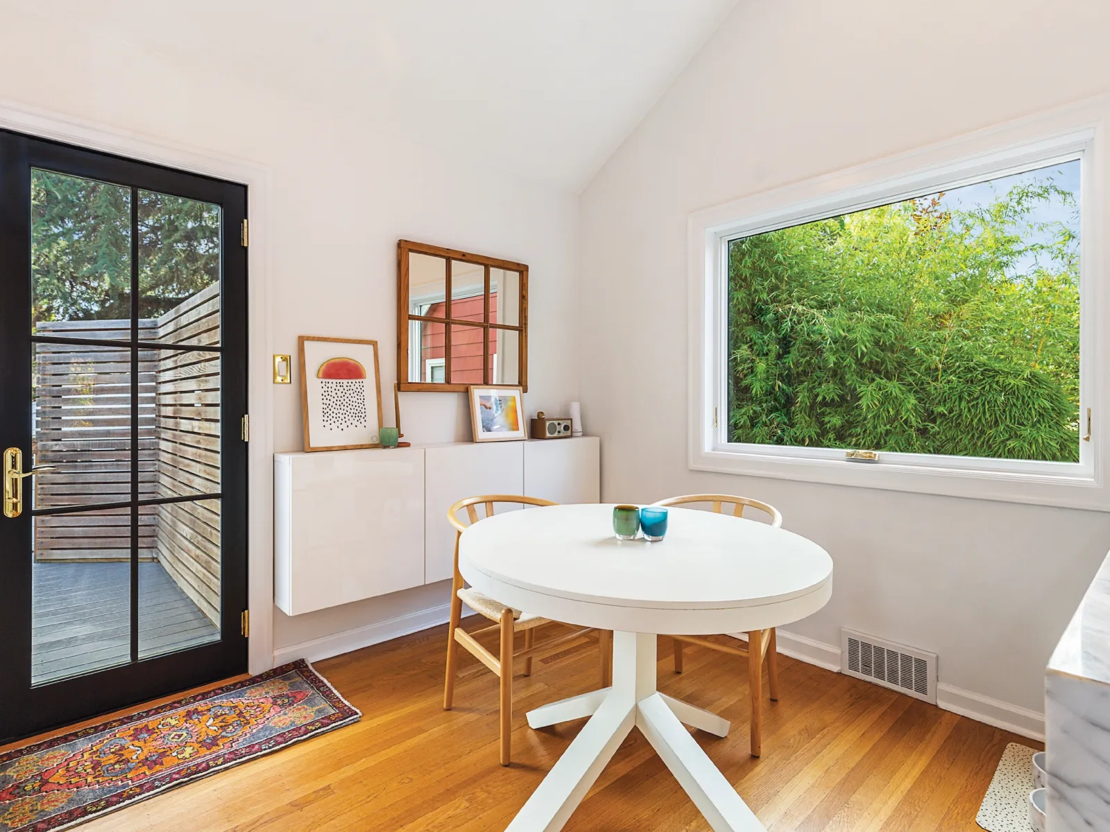 Bright dining area with round white table, wooden chairs, large window overlooking greenery, and black-framed glass door.