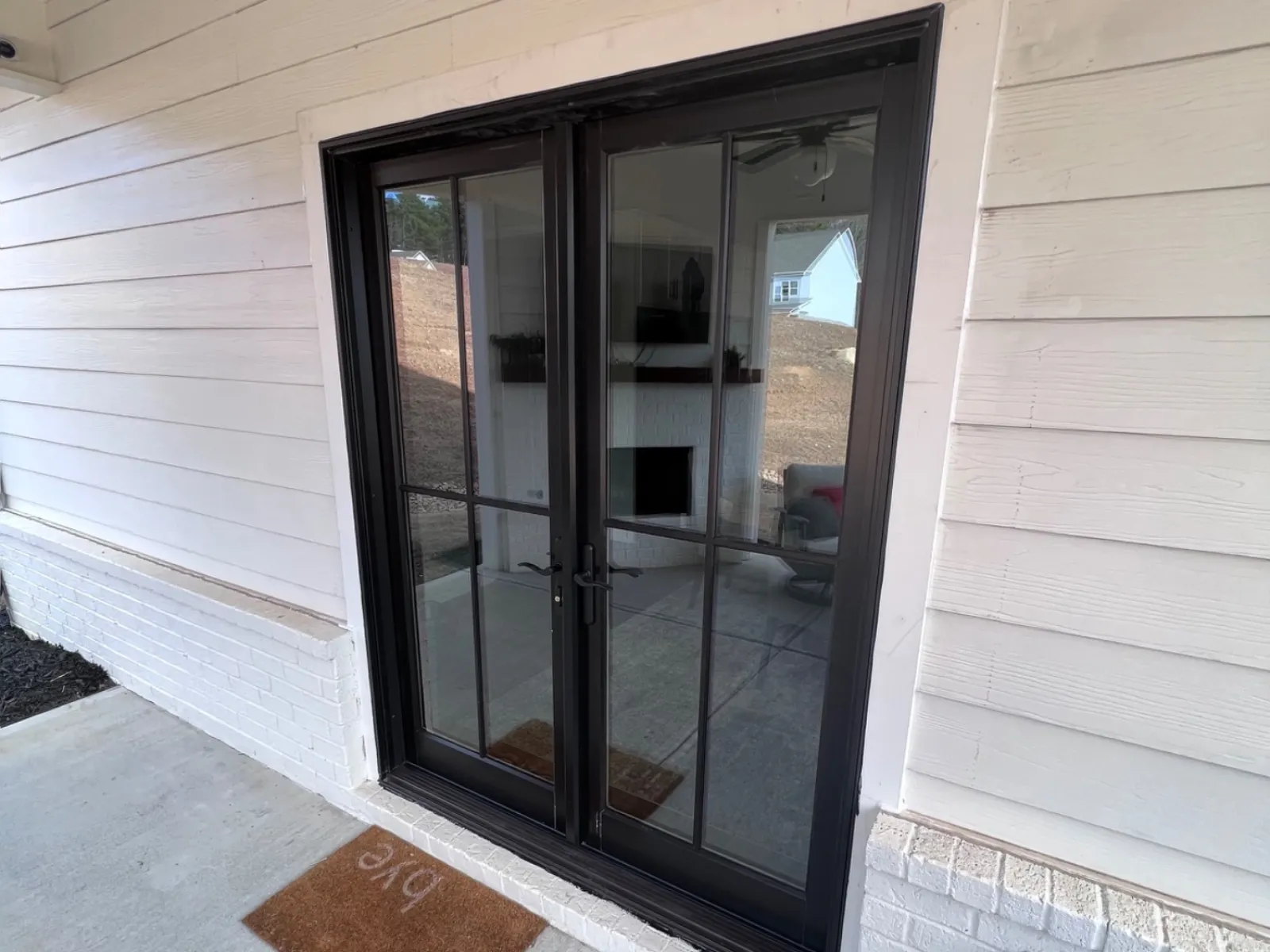 Black framed glass double doors on white house exterior with a brown doormat saying ‘hi’ on concrete porch.