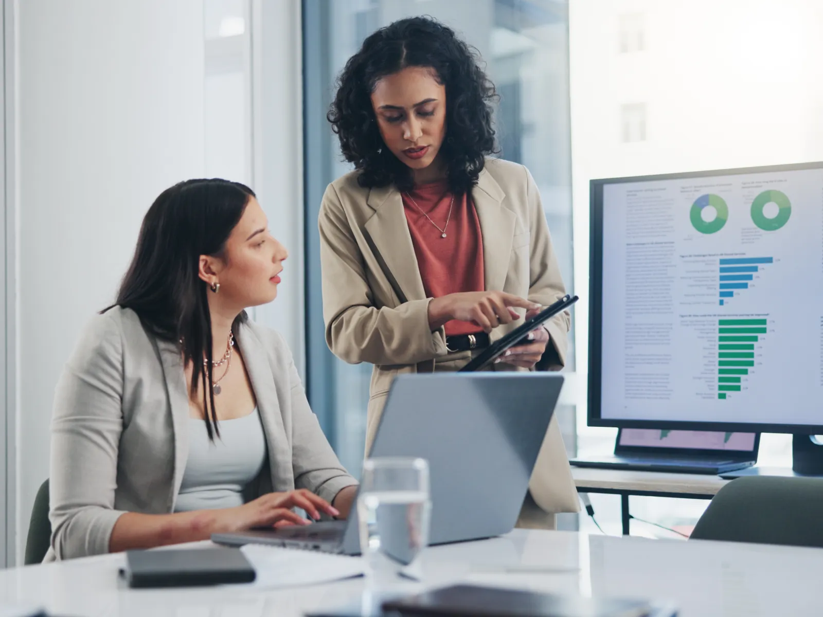 Two professional women discussing data charts on a tablet and computer in a modern office setting