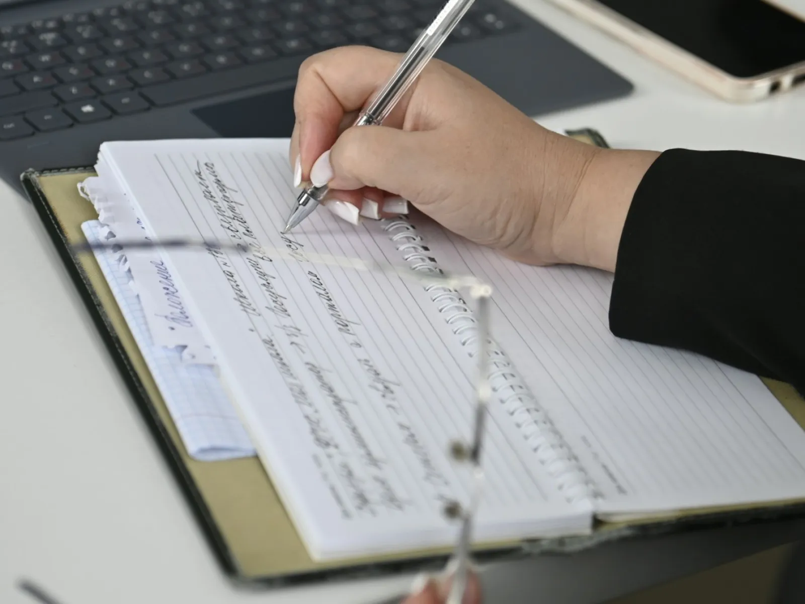 Person writing notes in spiral notebook with pen beside laptop and smartphone on desk