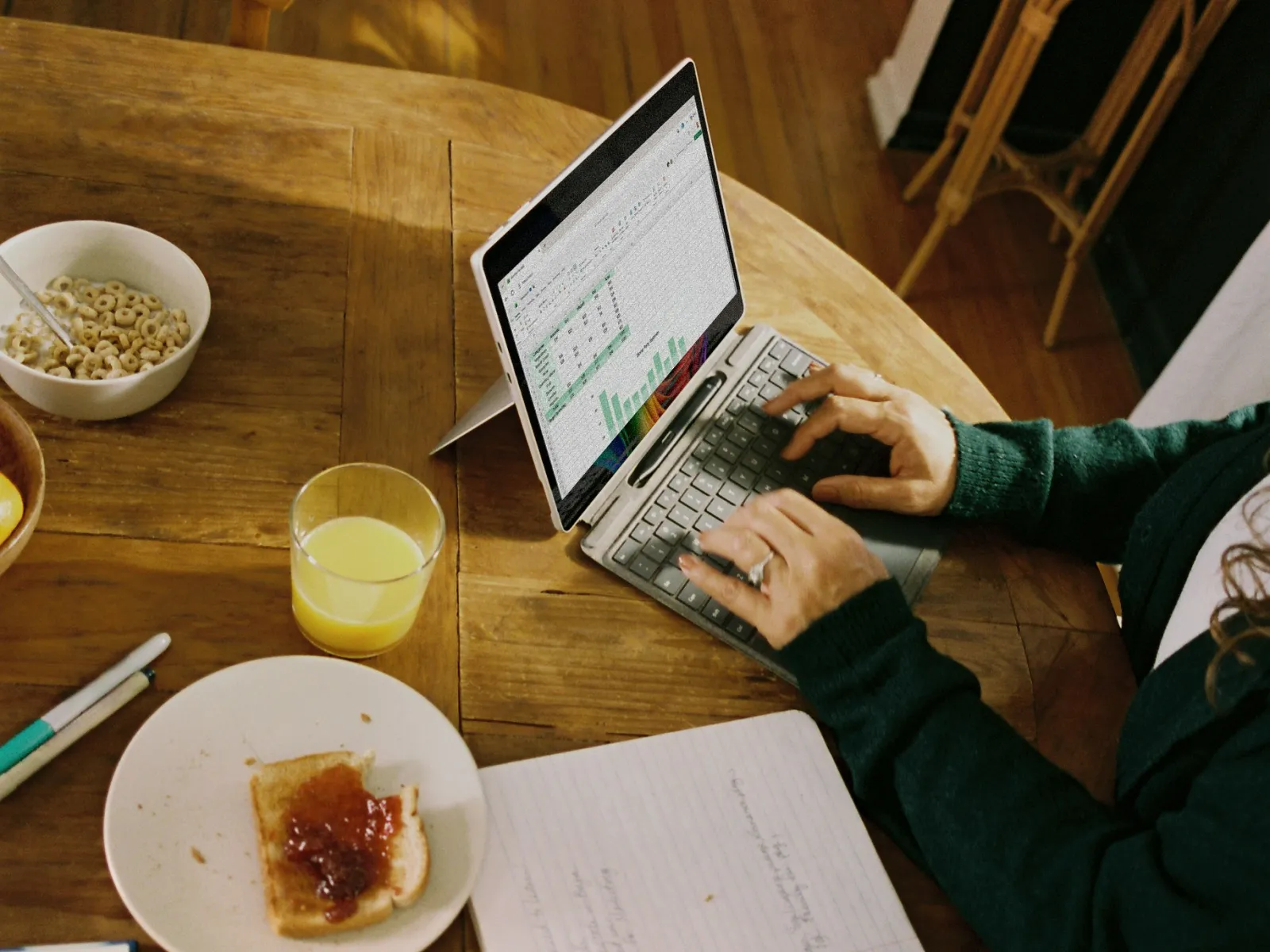 Person working on a tablet with keyboard at a dining table with breakfast, notebook, and smartphone.
