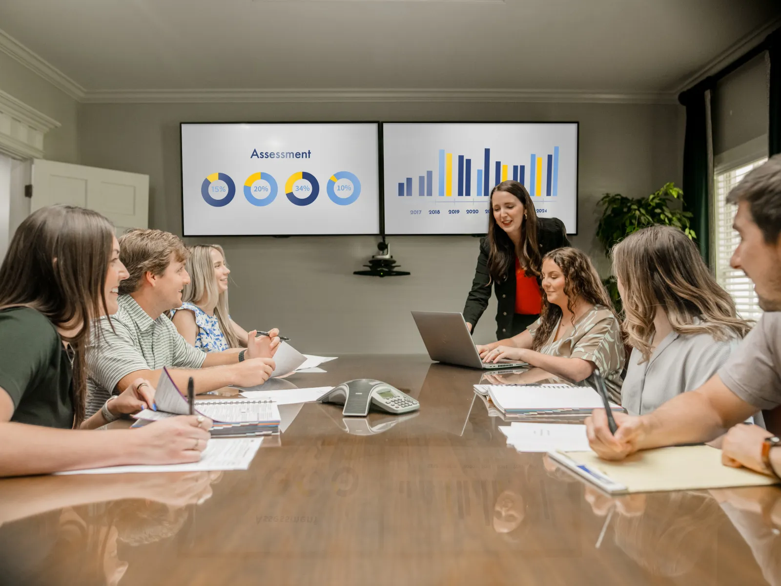 Team in a conference room discussing charts and graphs displayed on dual monitors during a business meeting