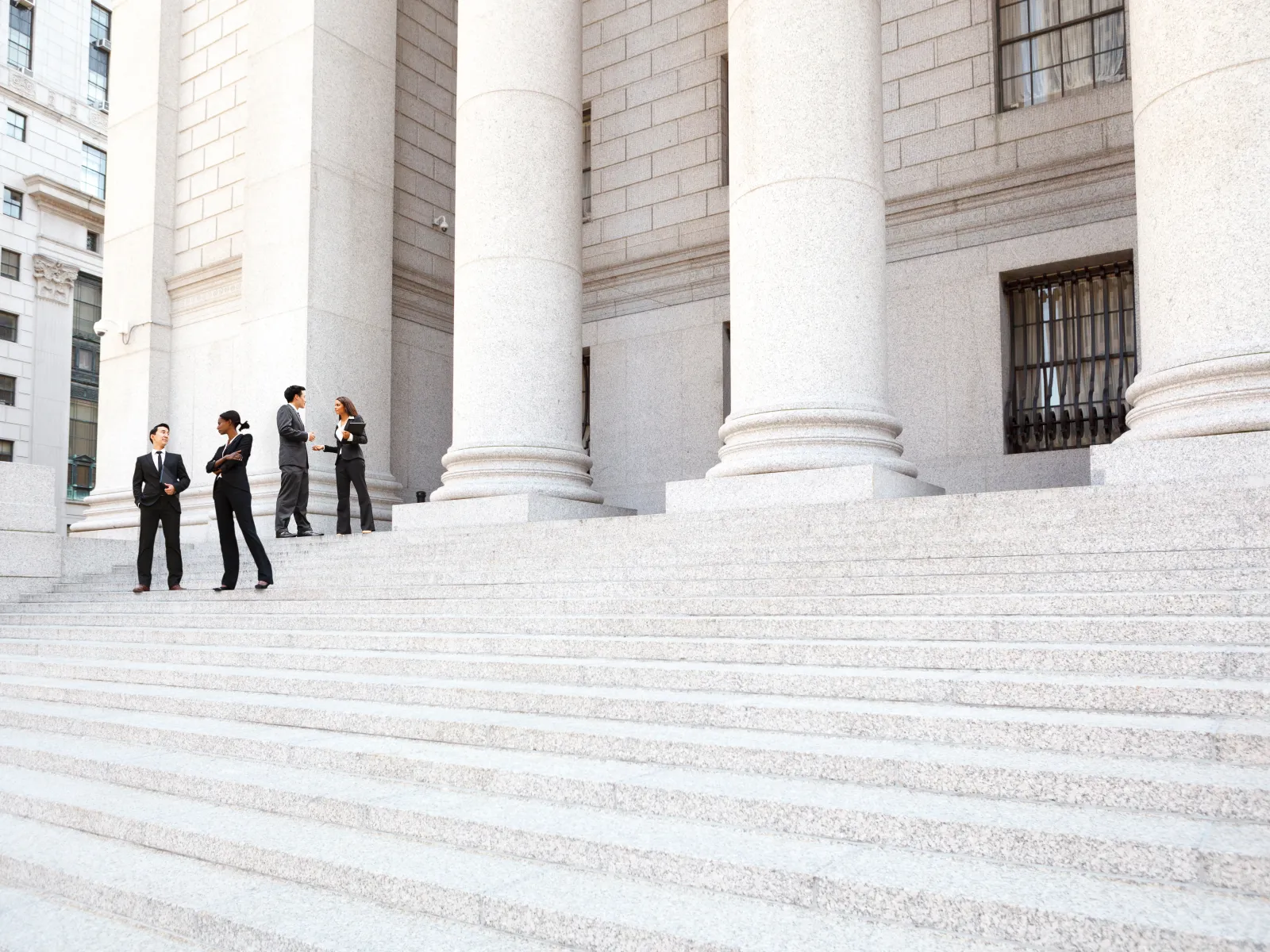 Business professionals in suits standing on large stone steps outside a government or courthouse building with tall columns.