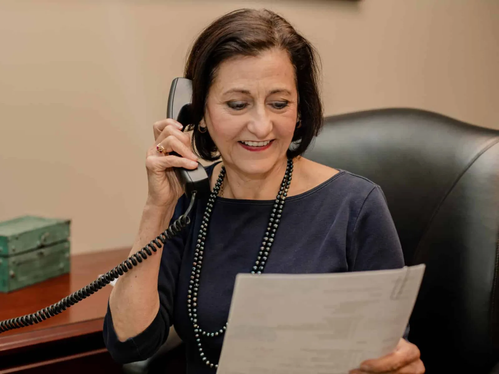 Middle-aged woman sitting at desk talking on phone while reading a document in office setting