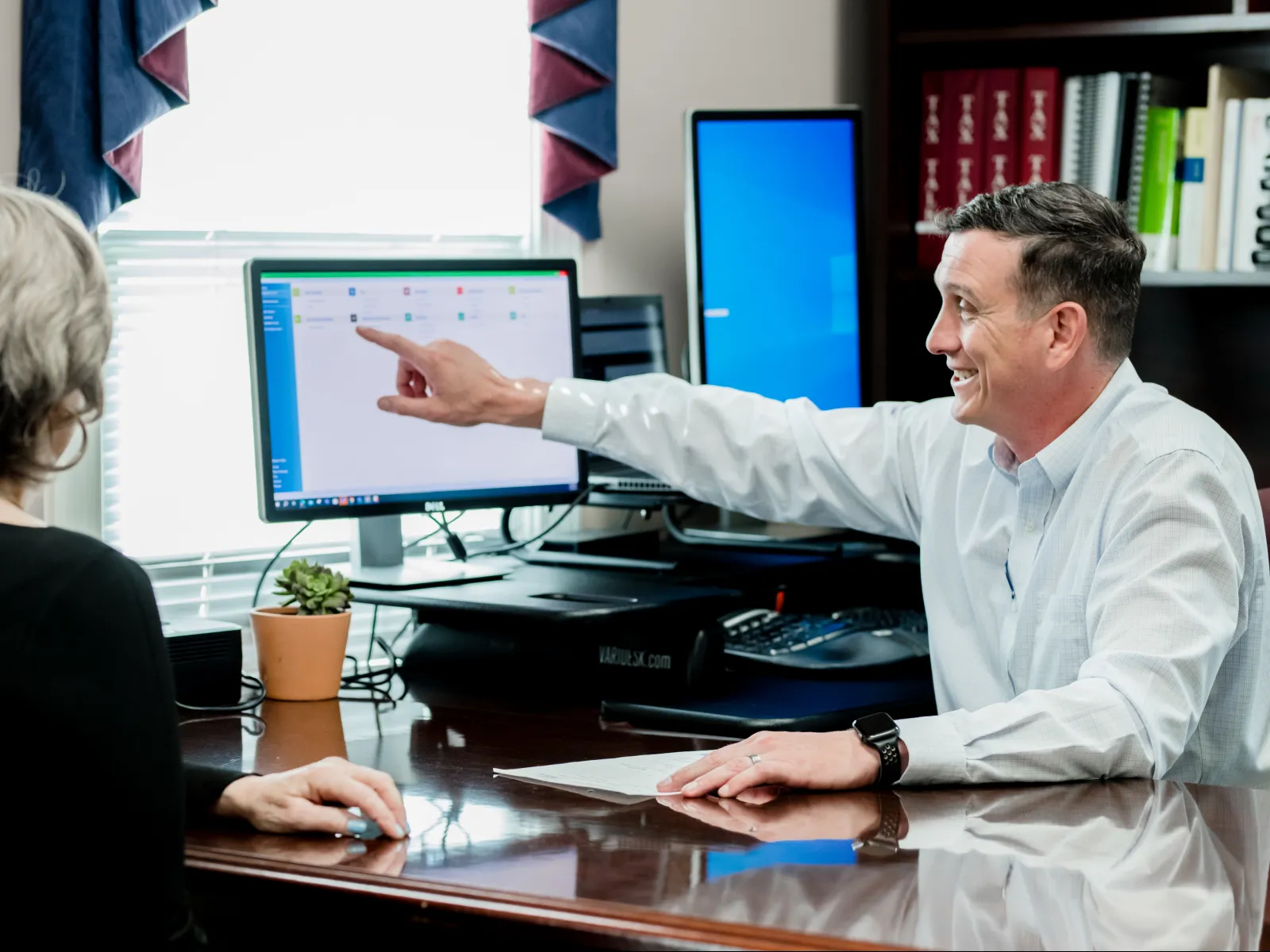 A man in a white shirt points at a computer screen while discussing with a woman in an office setting.