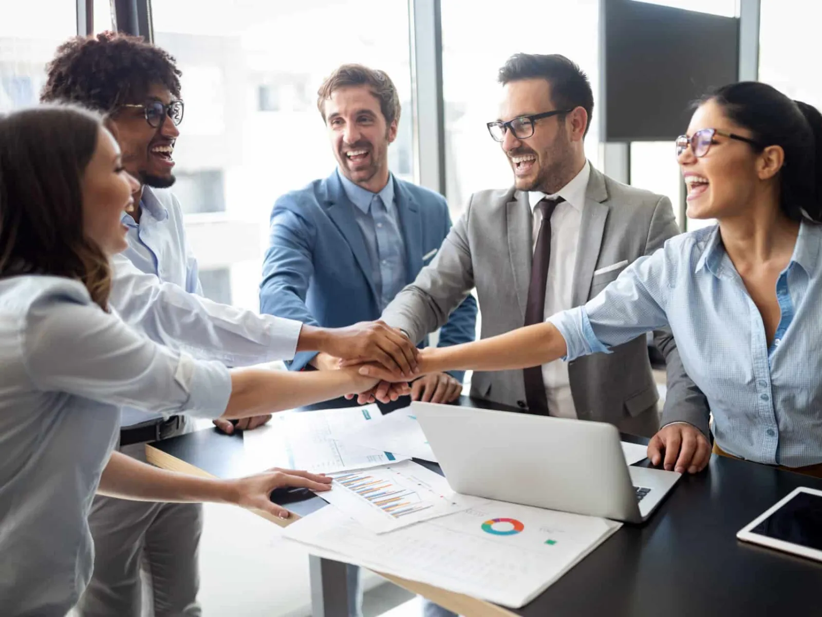 Diverse business team joining hands in office meeting for teamwork and success with charts and laptop