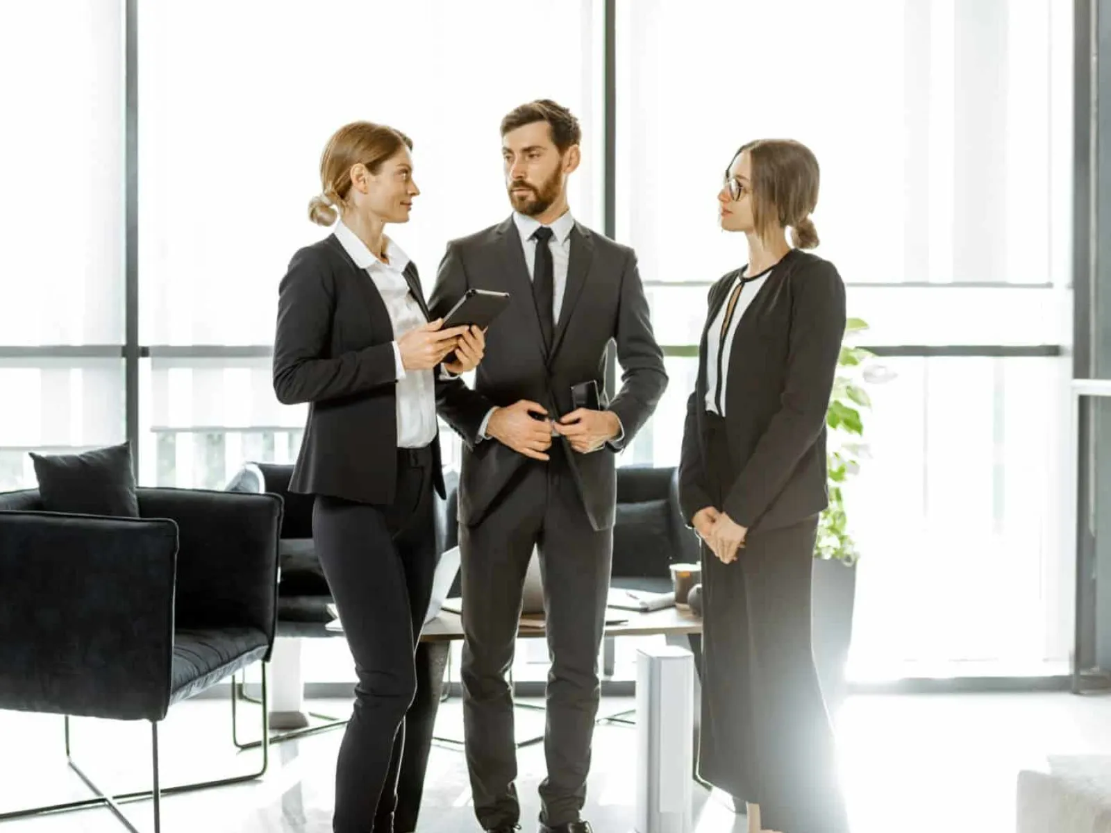 Three professionals in business attire standing and discussing in a modern office with large windows and minimal furniture