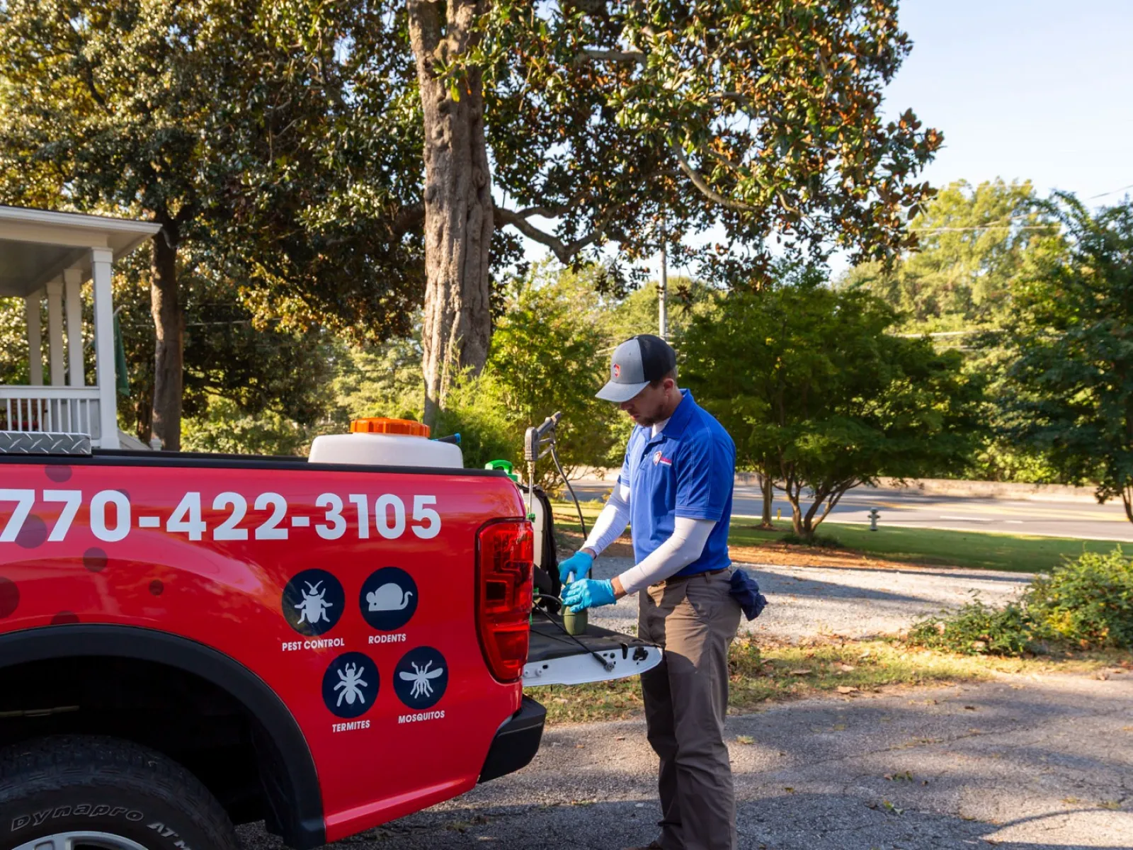 Pest control worker preparing equipment at the back of a red service truck in a residential neighborhood.