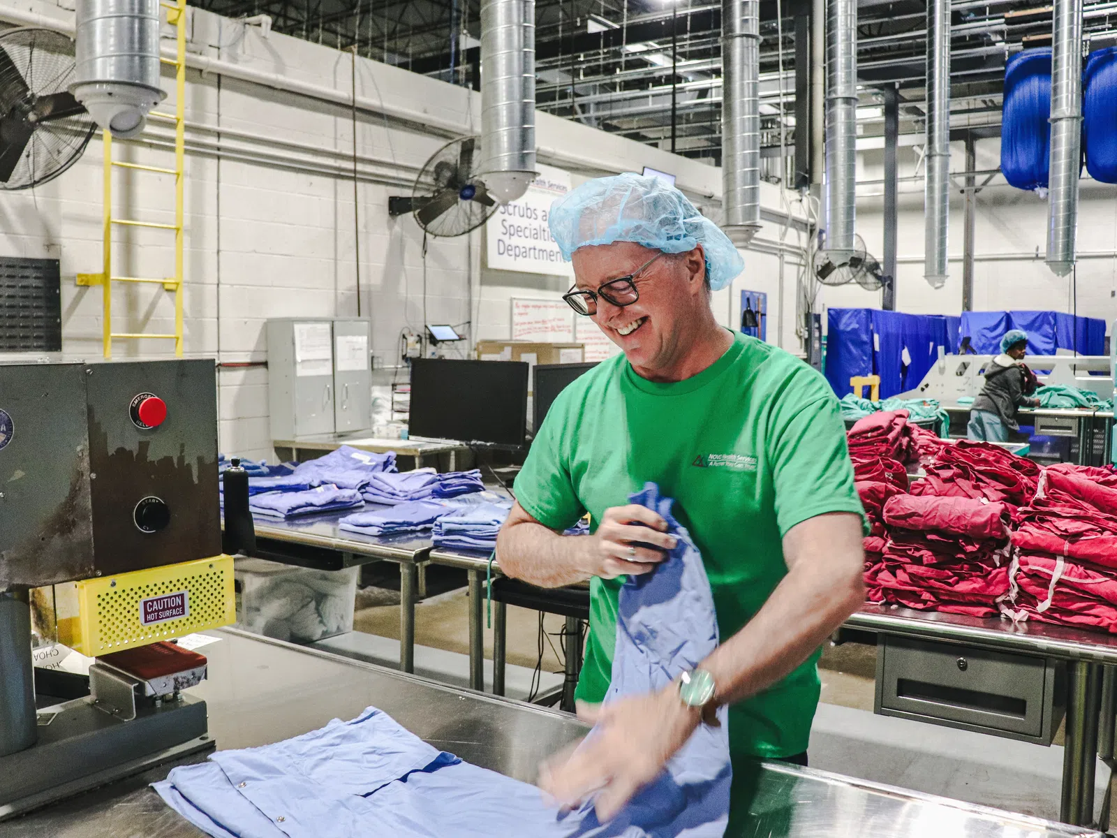 Smiling factory worker in green shirt sorting blue and red garments inside a large industrial facility.