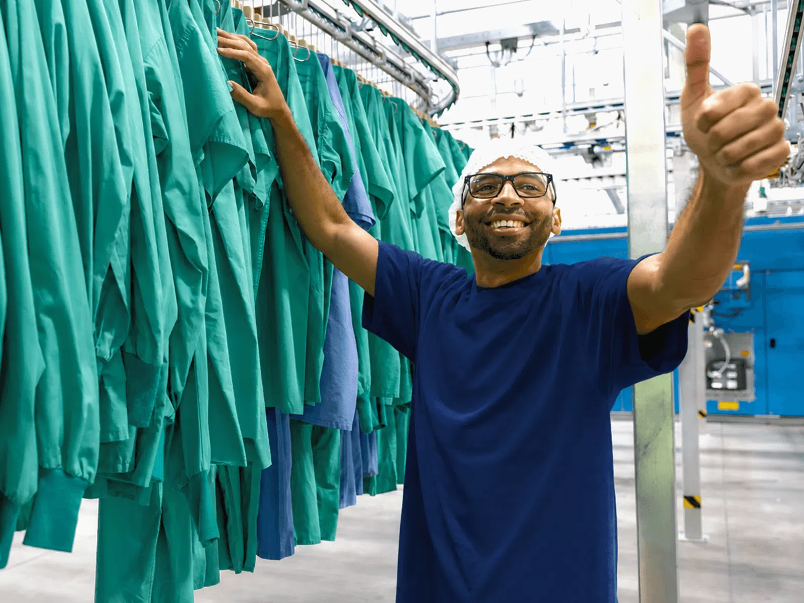 Smiling man in glasses gives thumbs up near hanging green and blue medical scrubs in industrial laundry.