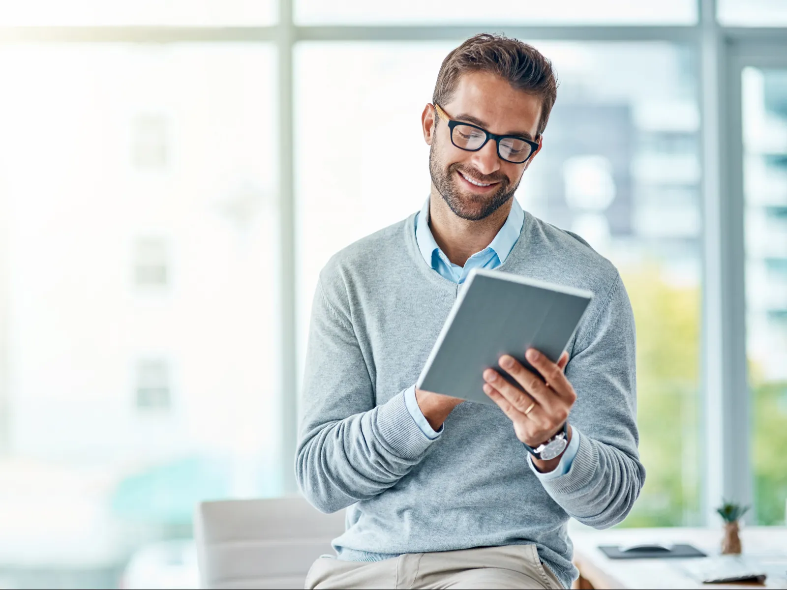 Smiling man wearing glasses and a gray sweater using a digital tablet in a bright modern office space