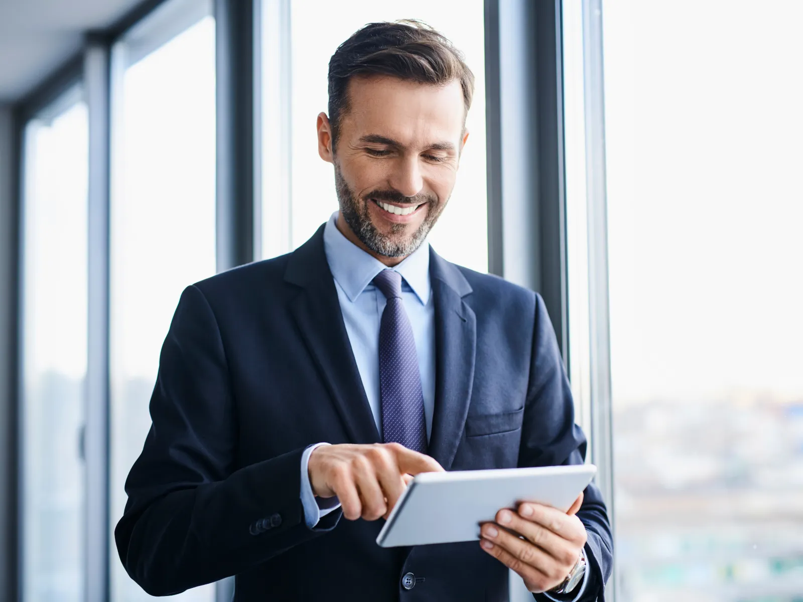 Smiling businessman in suit using tablet near office windows with city view in background