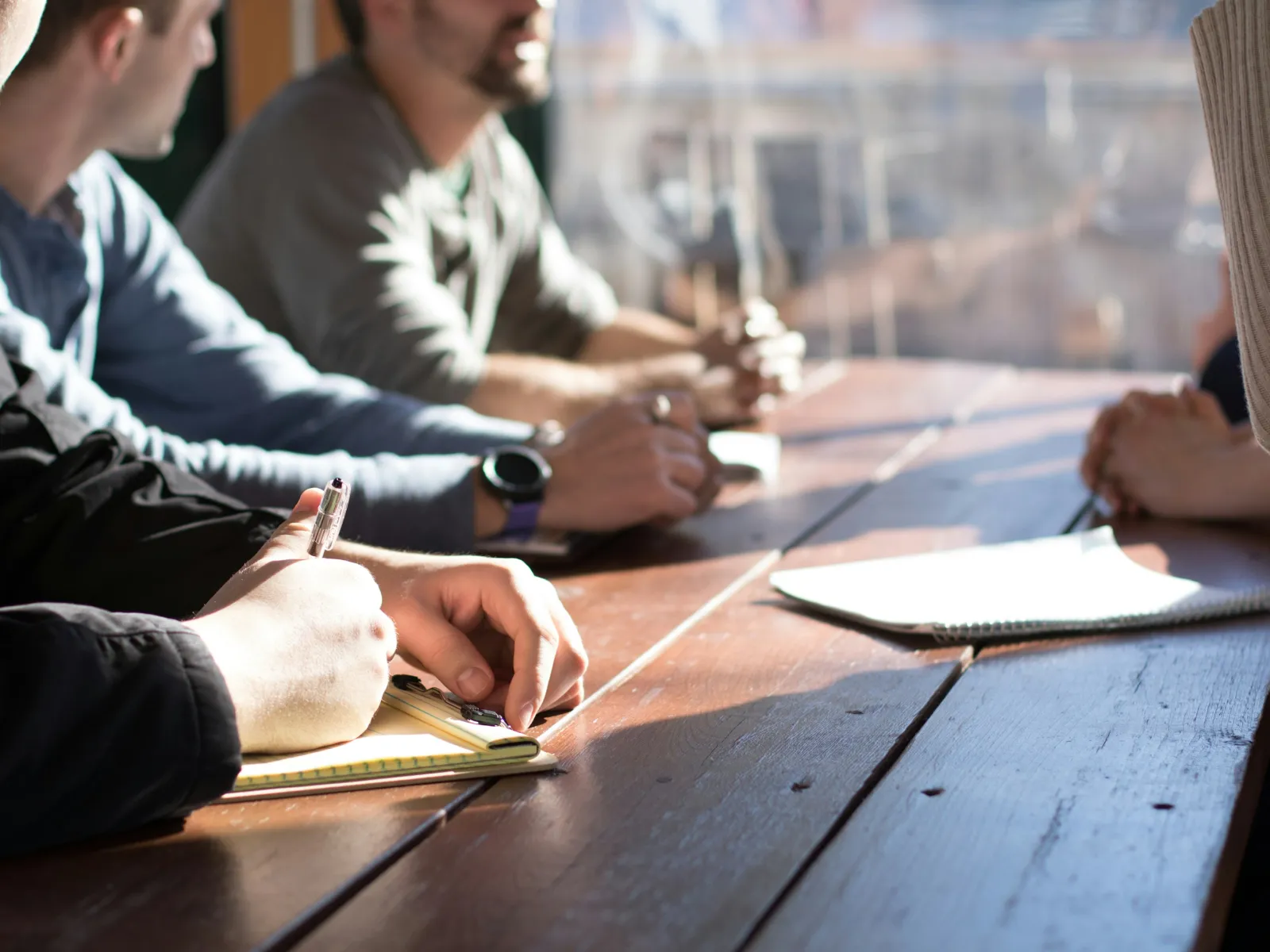 People sitting around a wooden table engaged in discussion and taking notes during a meeting in daylight.
