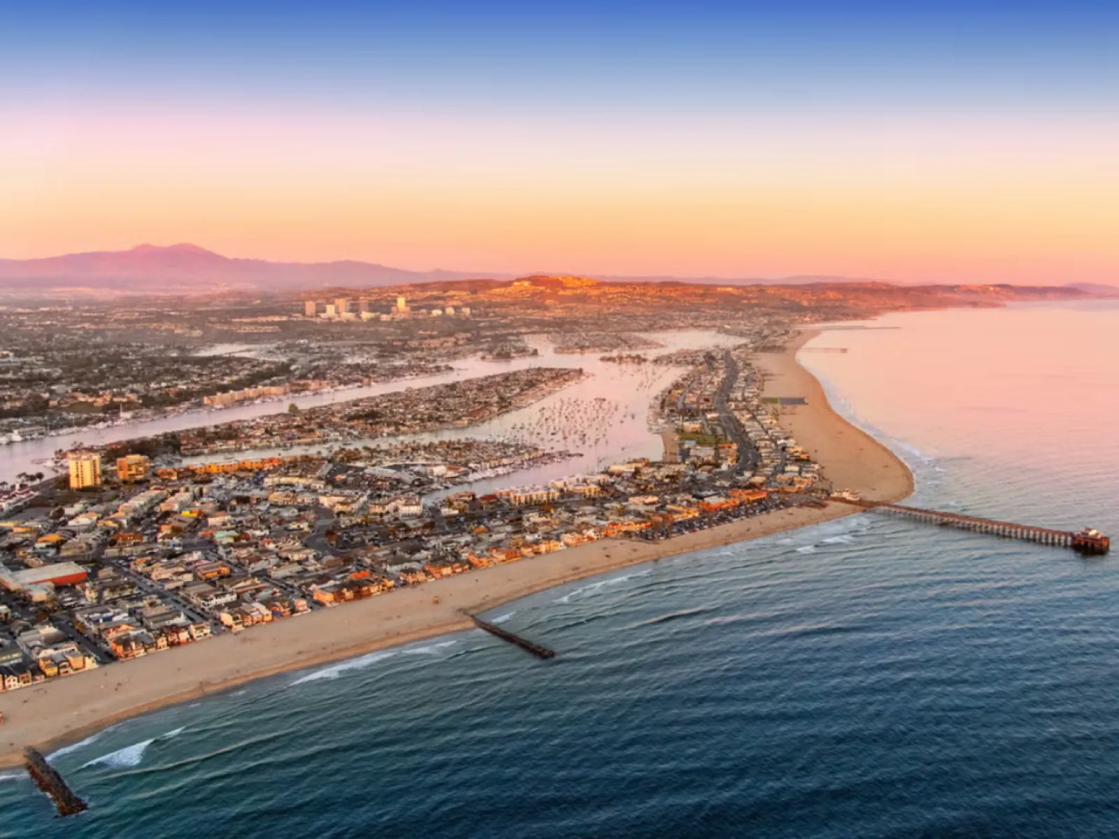Aerial view of a coastal city at sunset with a pier, sandy beach, and calm ocean waters extending to the horizon.