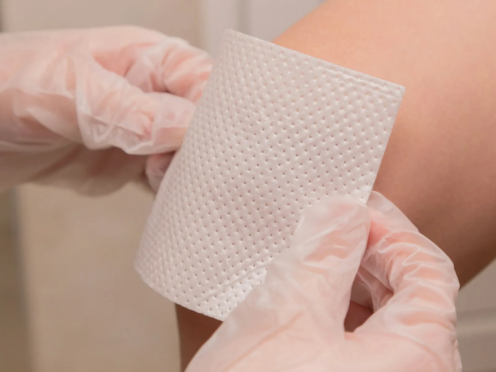 Hands in gloves applying a textured white adhesive patch on a person's bare knee for wound care or pain relief.