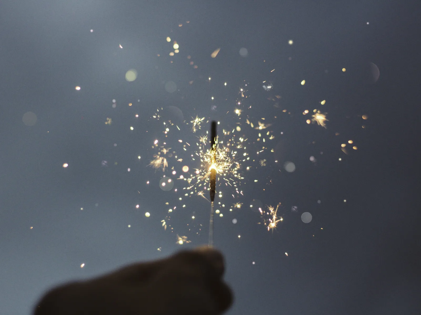Hand holding a sparkler emitting bright sparks against a dark gray background.