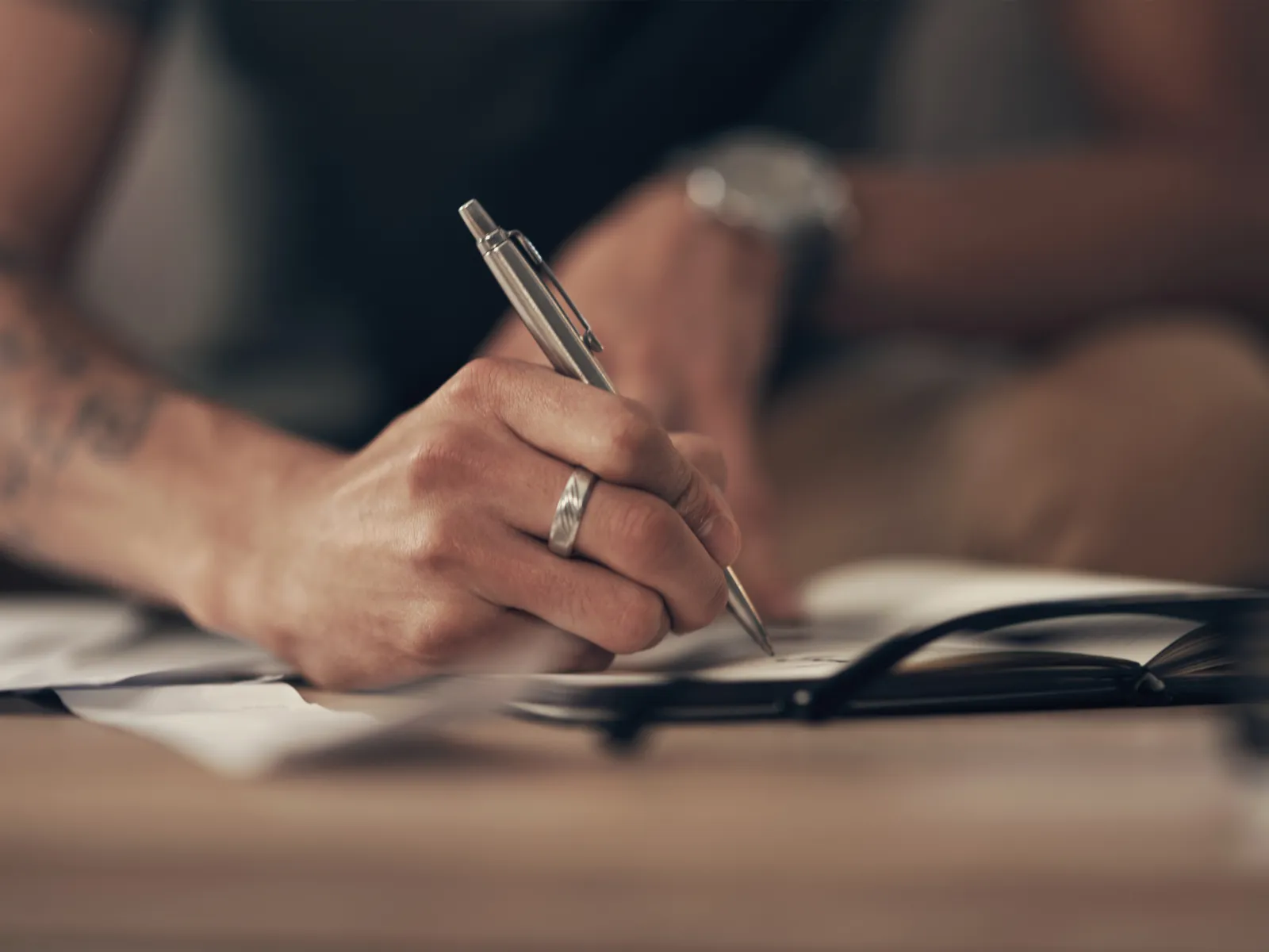Close-up of a tattooed hand with a ring writing with a pen on paper at a desk with eyeglasses nearby.