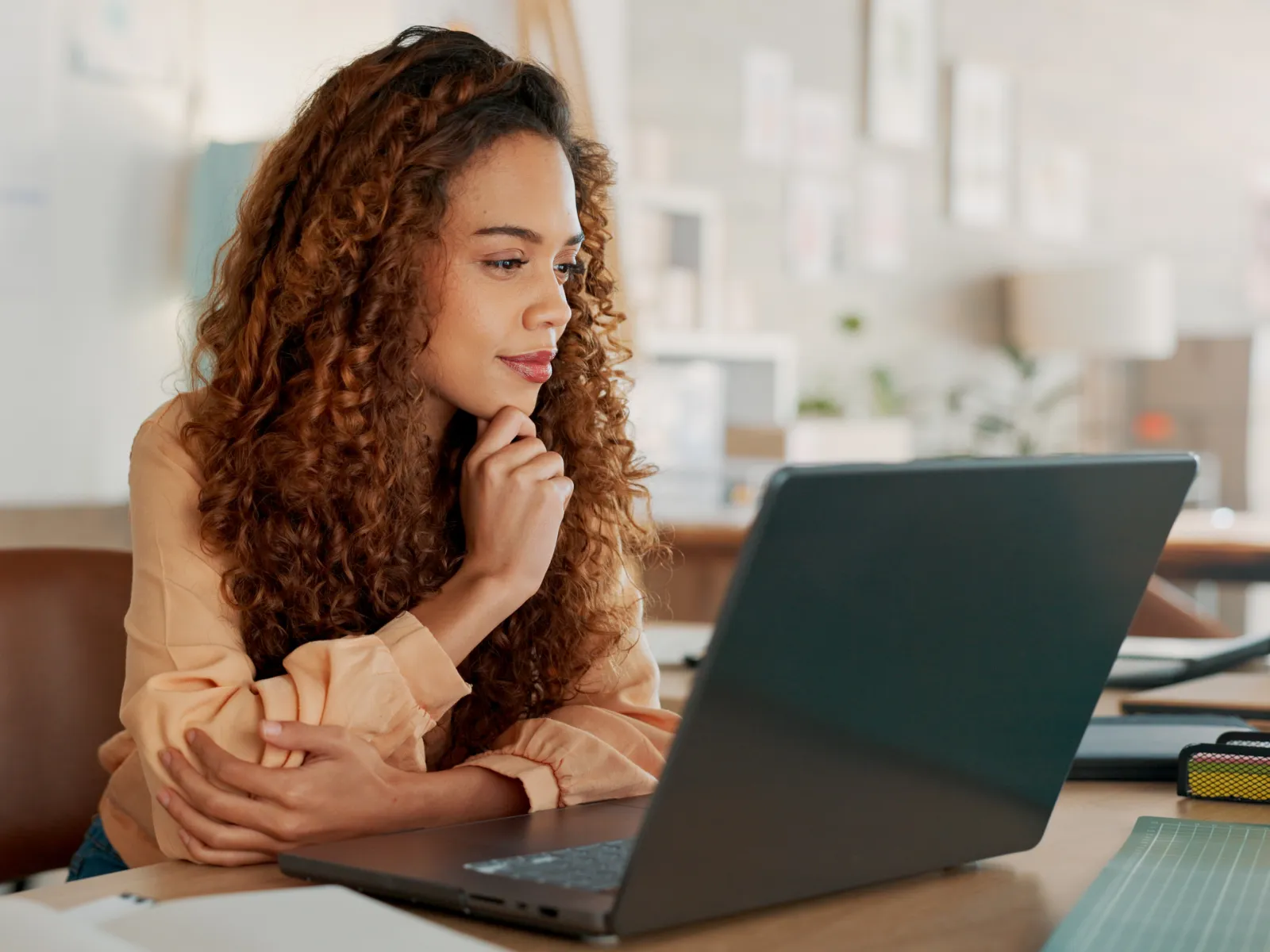 Young woman with curly hair focused on laptop while sitting at desk in bright modern office.