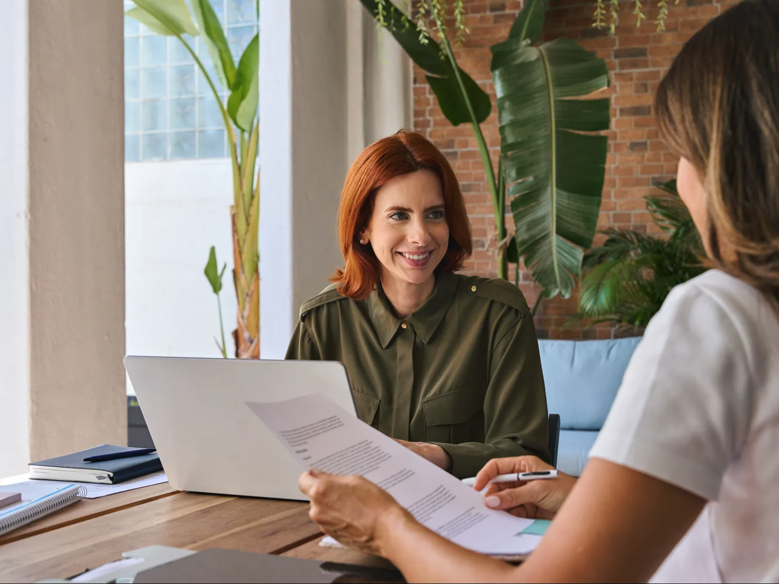 Two women discussing documents at a wooden table in a bright modern office with plants in the background