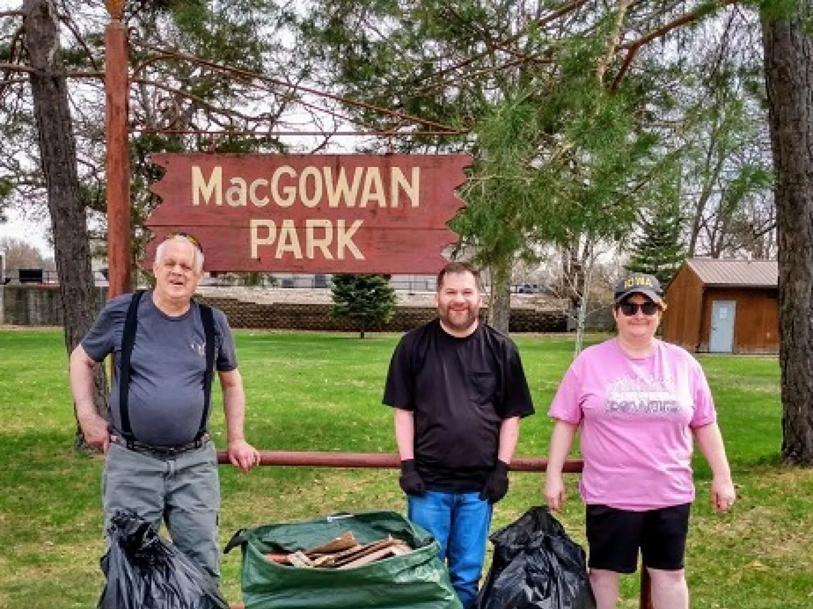 Three people standing in front of MacGowan Park sign with bags of collected trash during community cleanup.