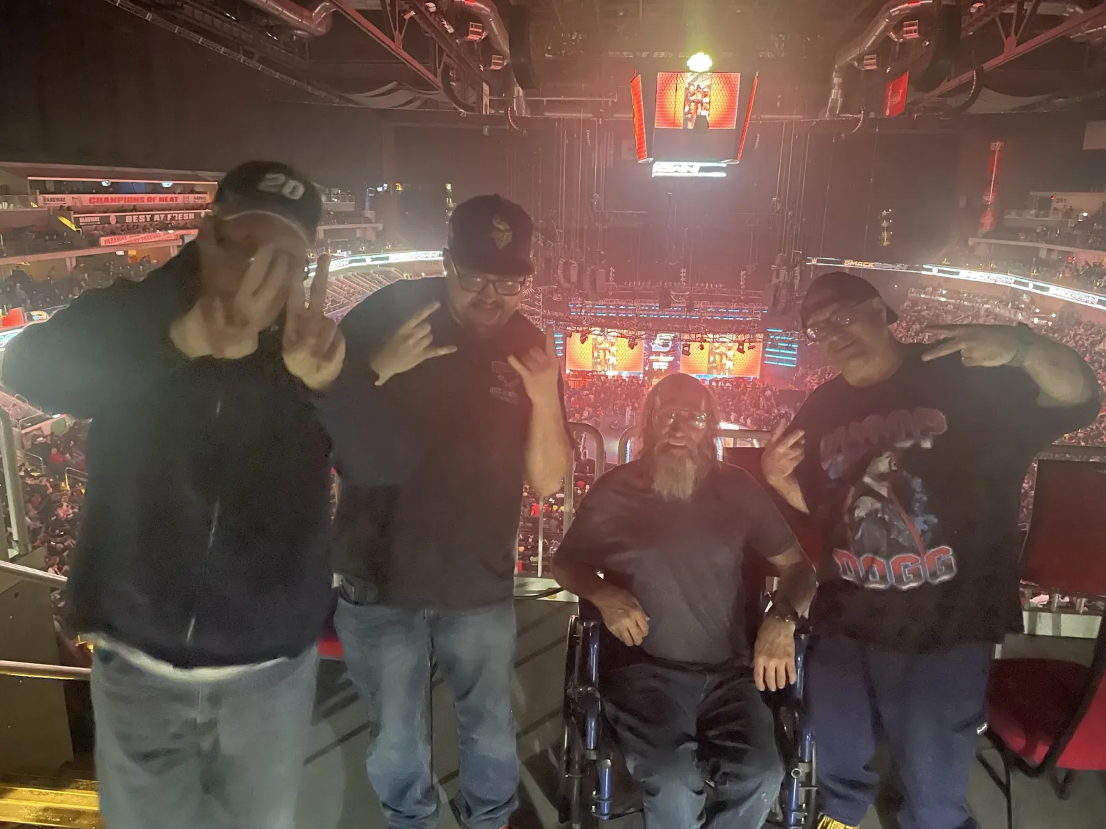 Four men posing with hand signs inside a crowded arena with a stage and screens in the background.
