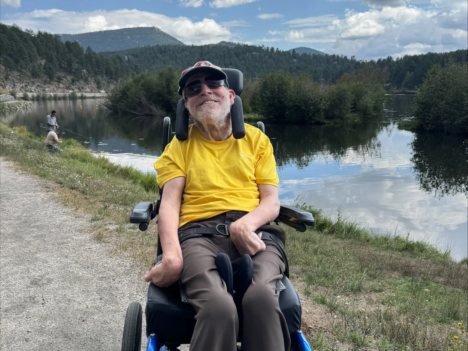 Smiling man in a yellow shirt sitting in a blue wheelchair by a lakeside with mountains and cloudy sky in the background