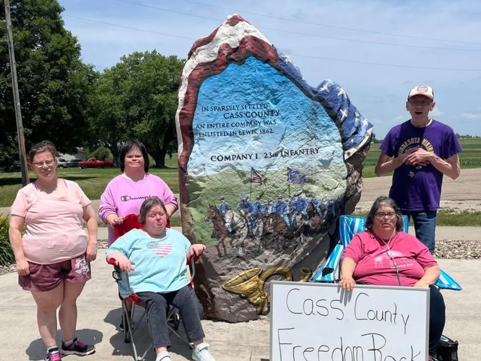 Group of five people posing near the painted Freedom Rock in Cass County with a commemorative sign