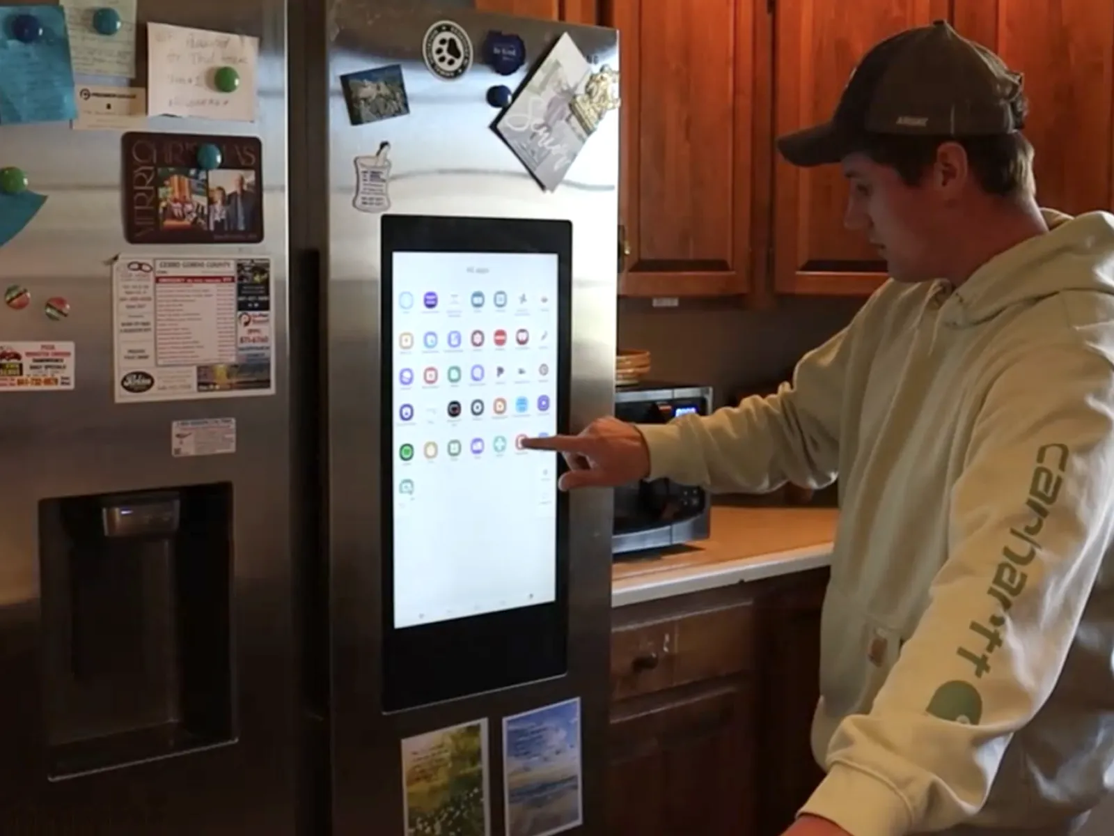 Person using smart refrigerator with touchscreen display in a wooden kitchen interior.