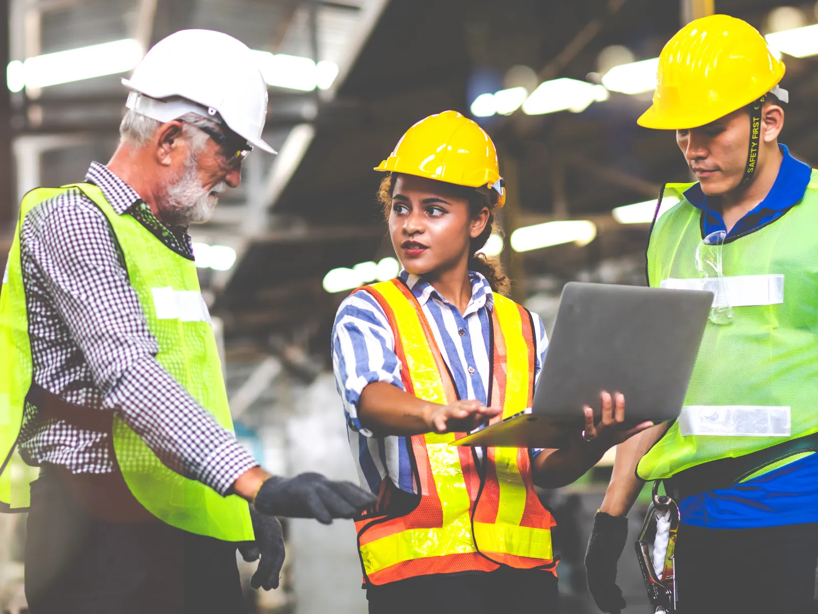 Three workers in hard hats and vests discussing plans with a laptop in a warehouse setting.