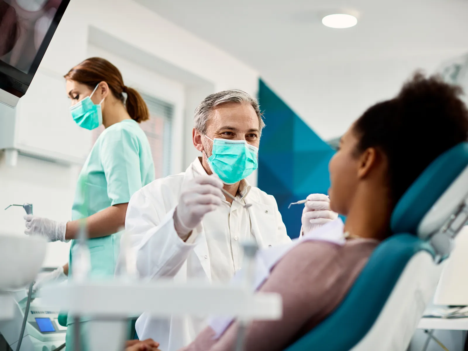 Dentist in mask consulting with patient in dental clinic, staff preparing in background.