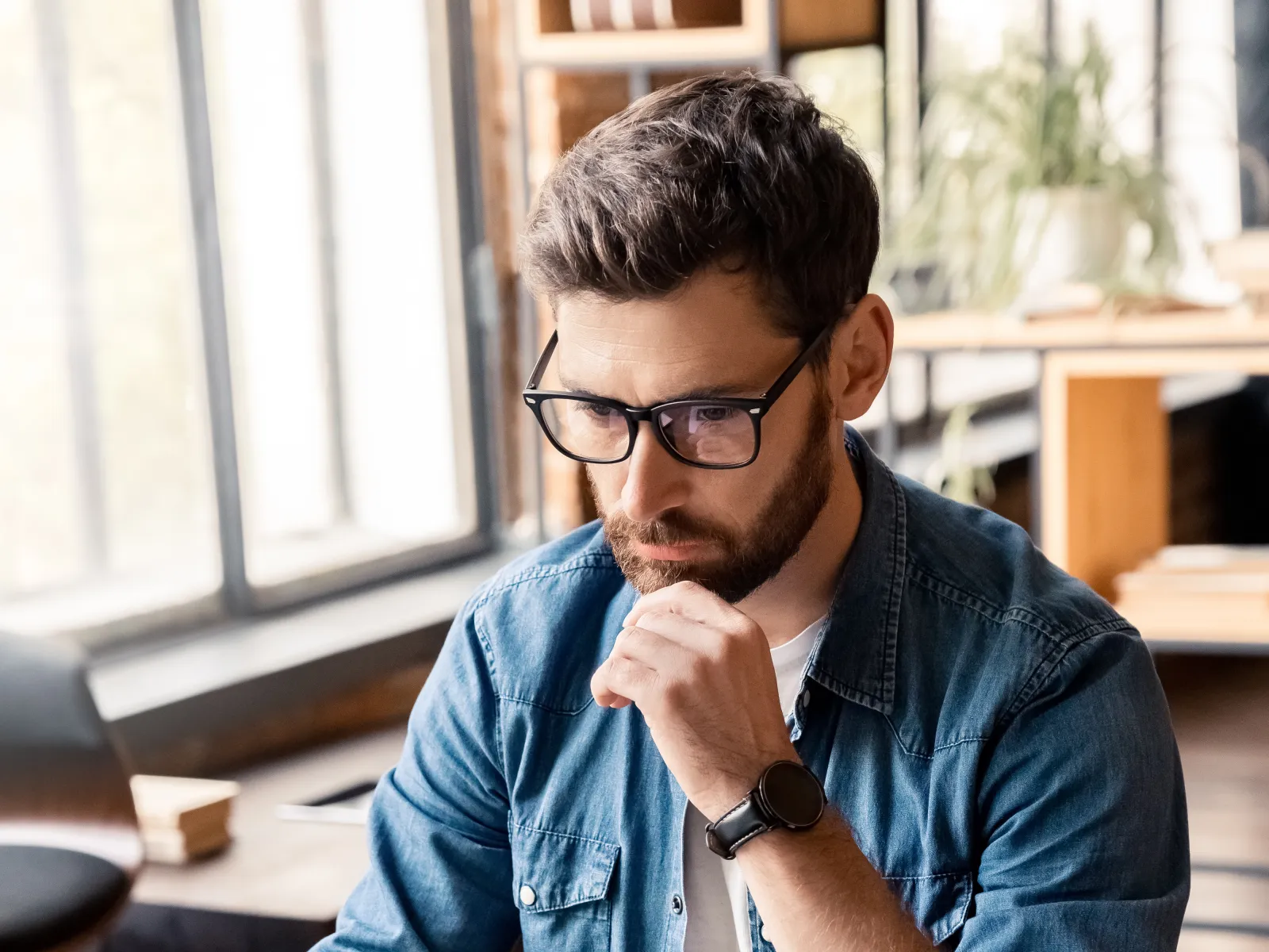 Young man with glasses working intently on a laptop in a modern office space.