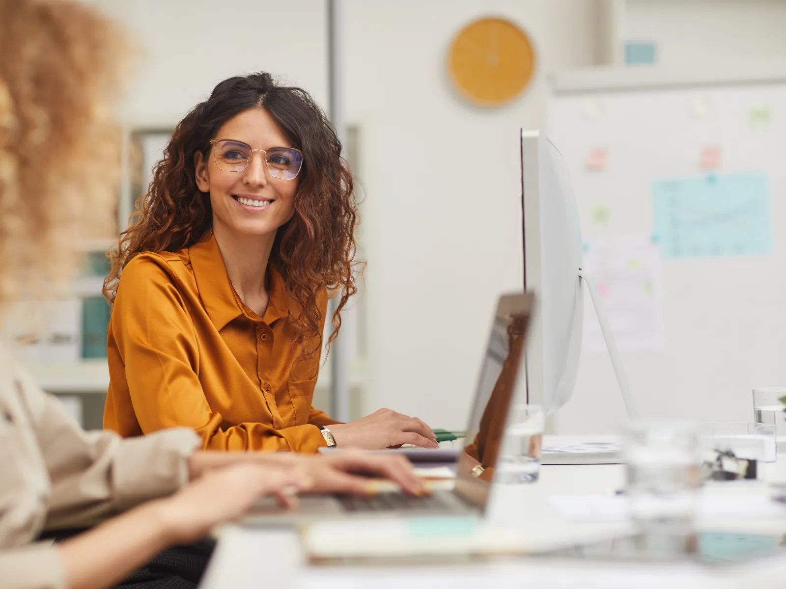 A woman in glasses smiles while working beside a colleague at a bright office desk.