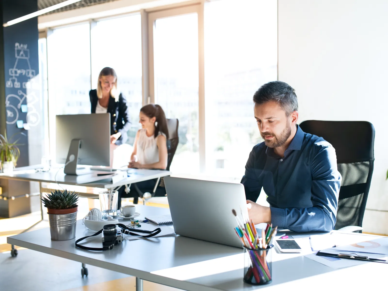 Man working on laptop at desk with office supplies in bright modern office with two women collaborating in background