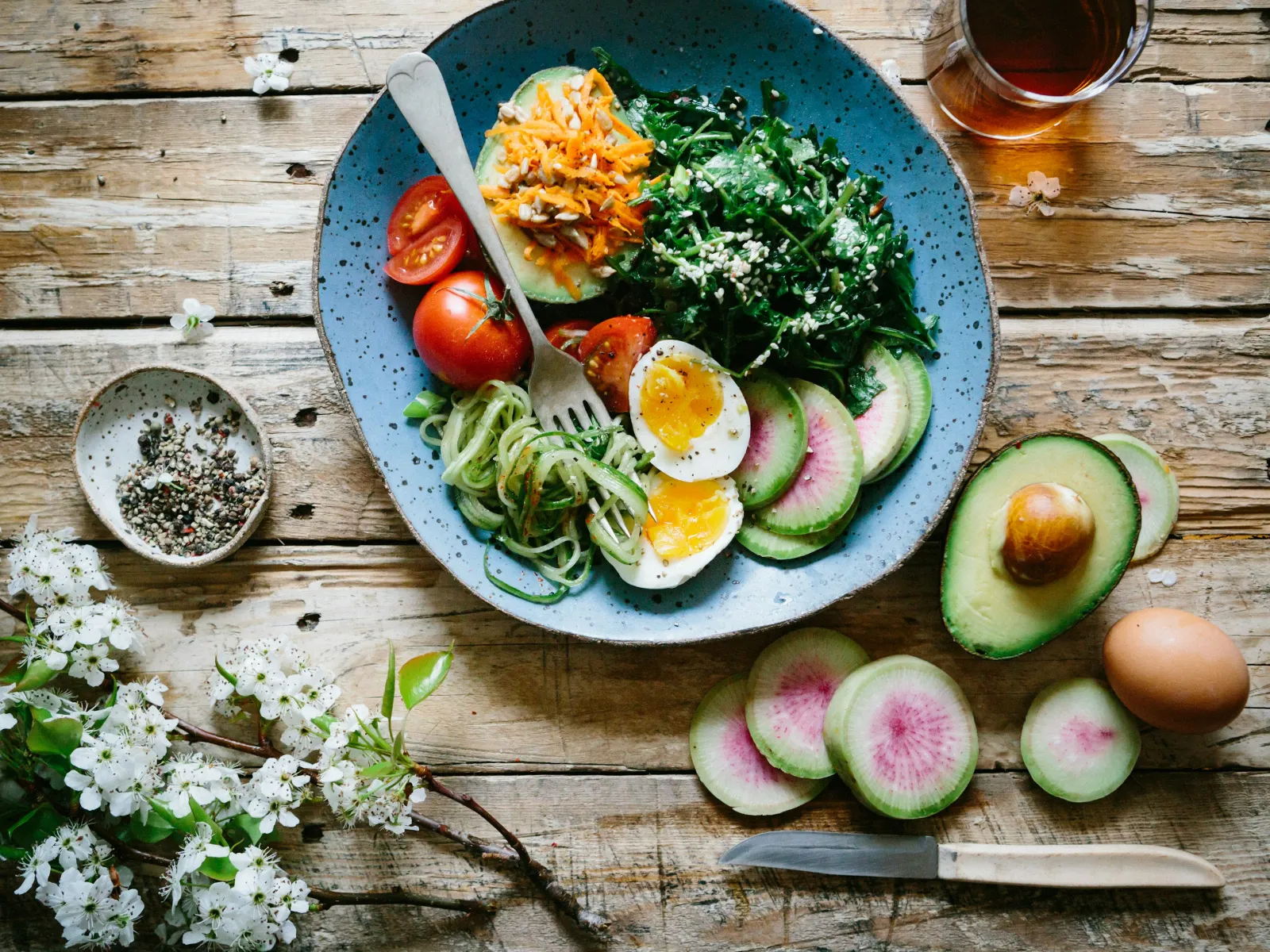 Healthy bowl with boiled eggs, avocado, tomatoes, greens, and spiralized zucchini on rustic wooden table.