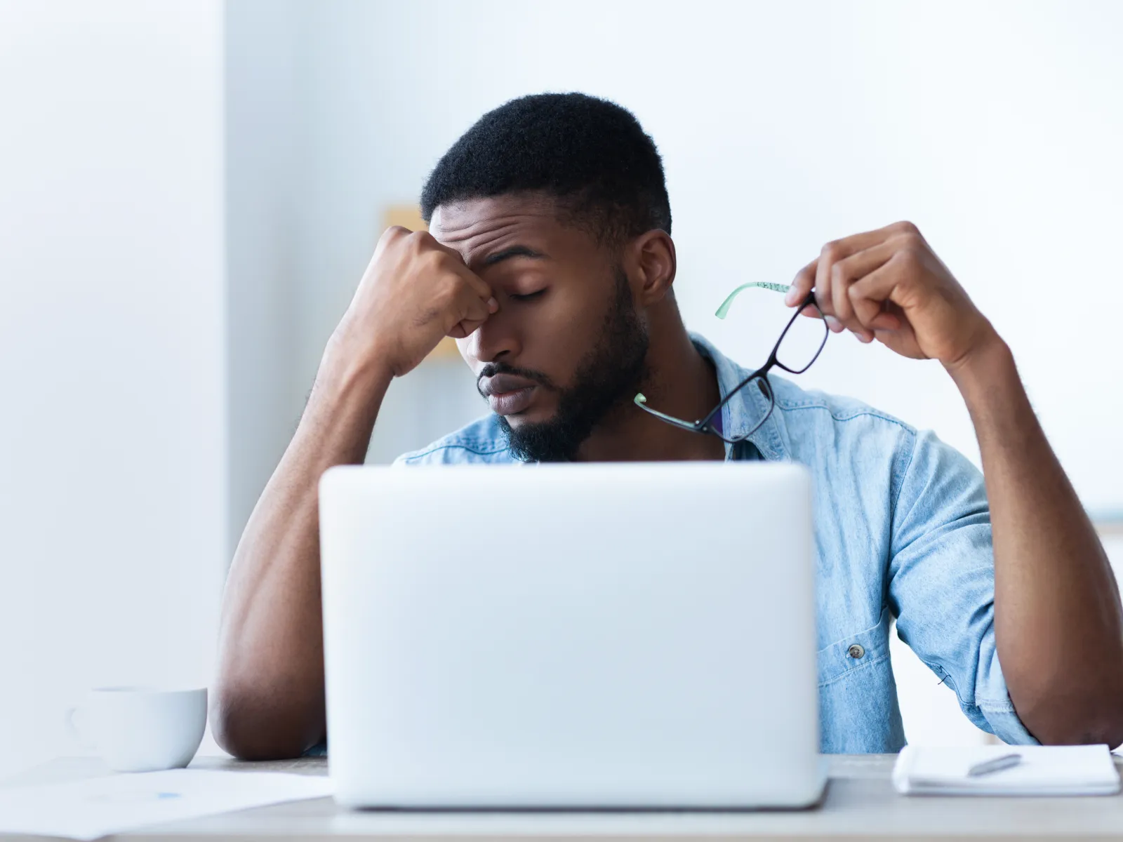 Tired young man rubbing eyes while holding glasses sitting in front of laptop at desk