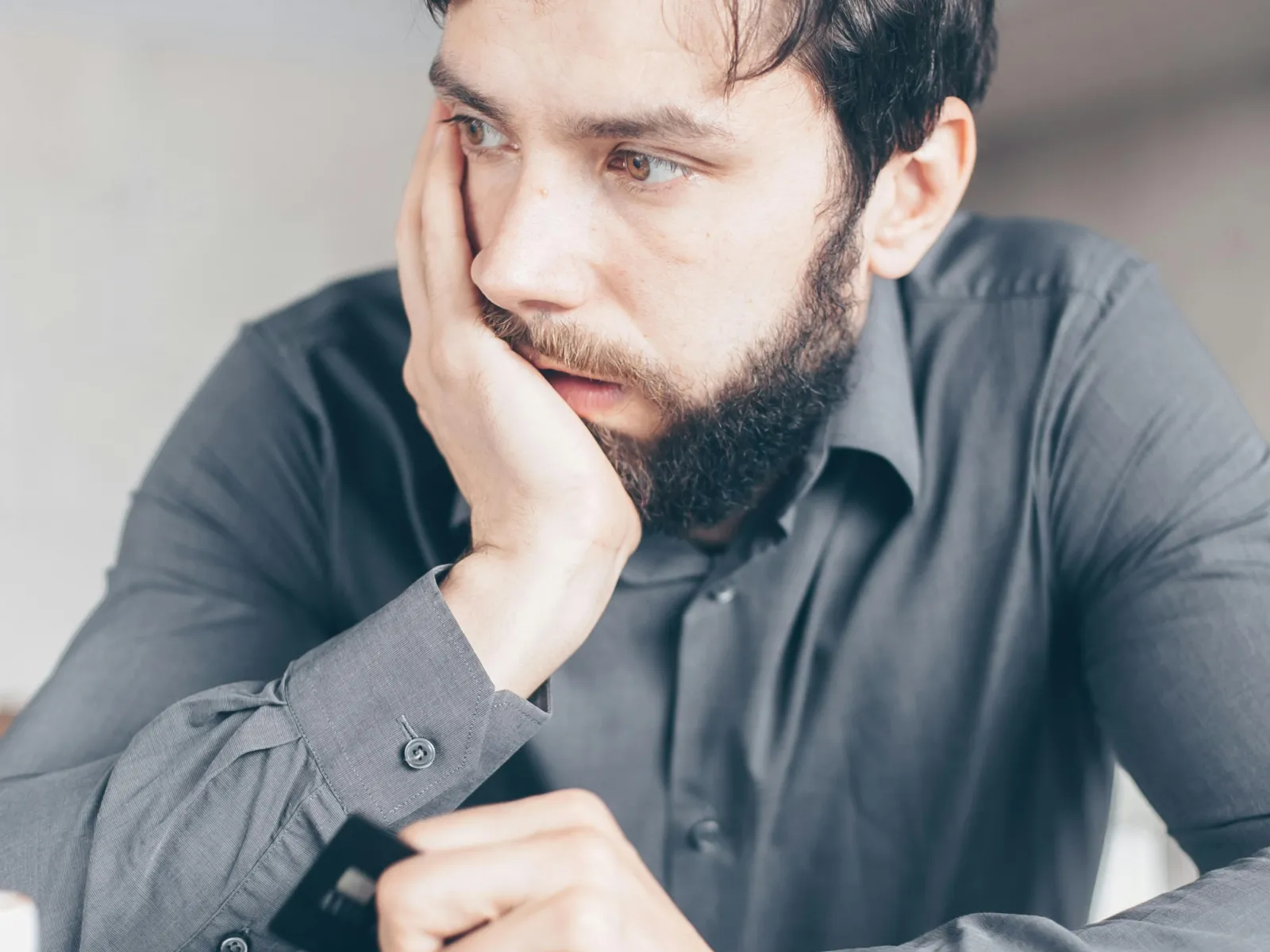 Thoughtful man holding a credit card, looking away while sitting at table with smartphone and glasses.