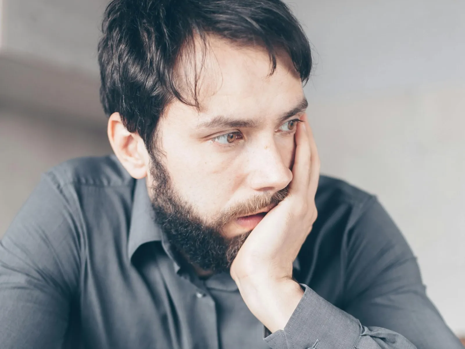 Bearded man in gray shirt looking worried holding a credit card with phone and glasses on table.